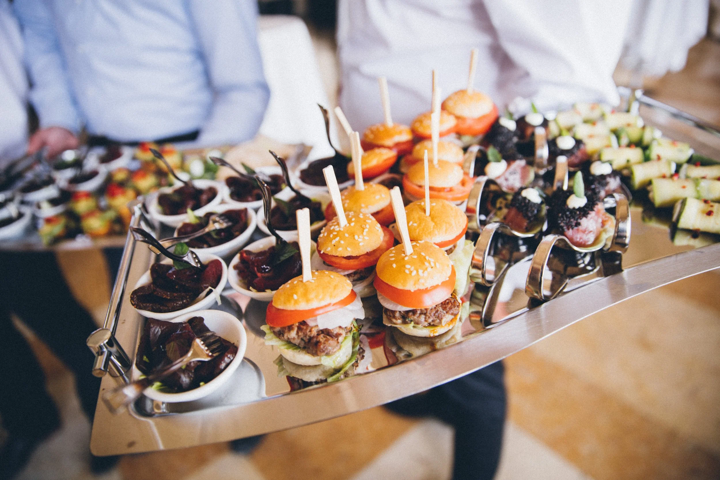 A tray of assorted mini sandwiches and appetizers, including sliders with buns, lettuce, tomato, and meat, as well as small cups of what appears to be desserts or dips, served at a buffet or party.