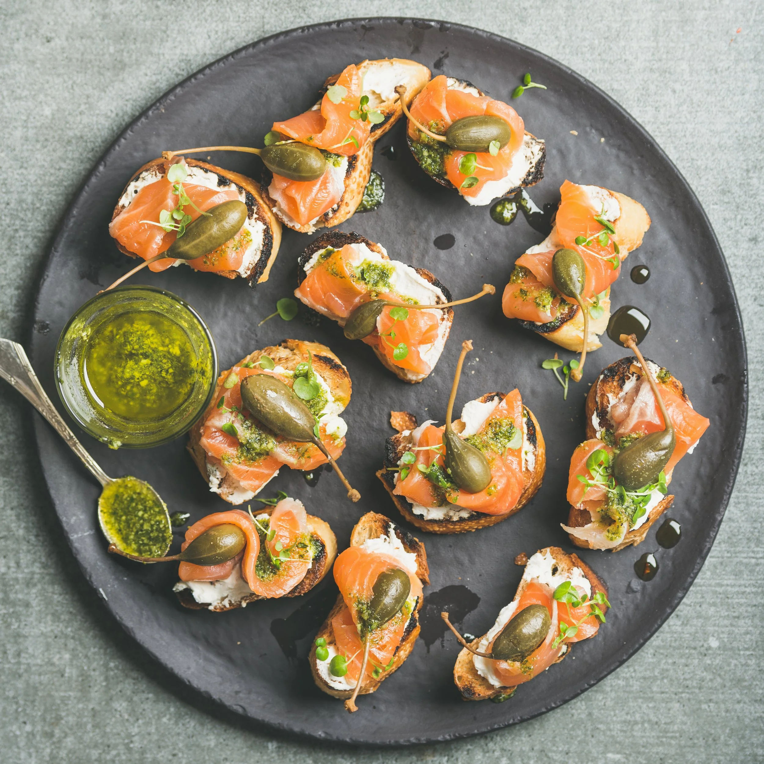 Sliced baguette topped with cream cheese, smoked salmon, capers, microgreens, and drizzled with green sauce, served on a black oval plate with a small bowl of green oil on the side.