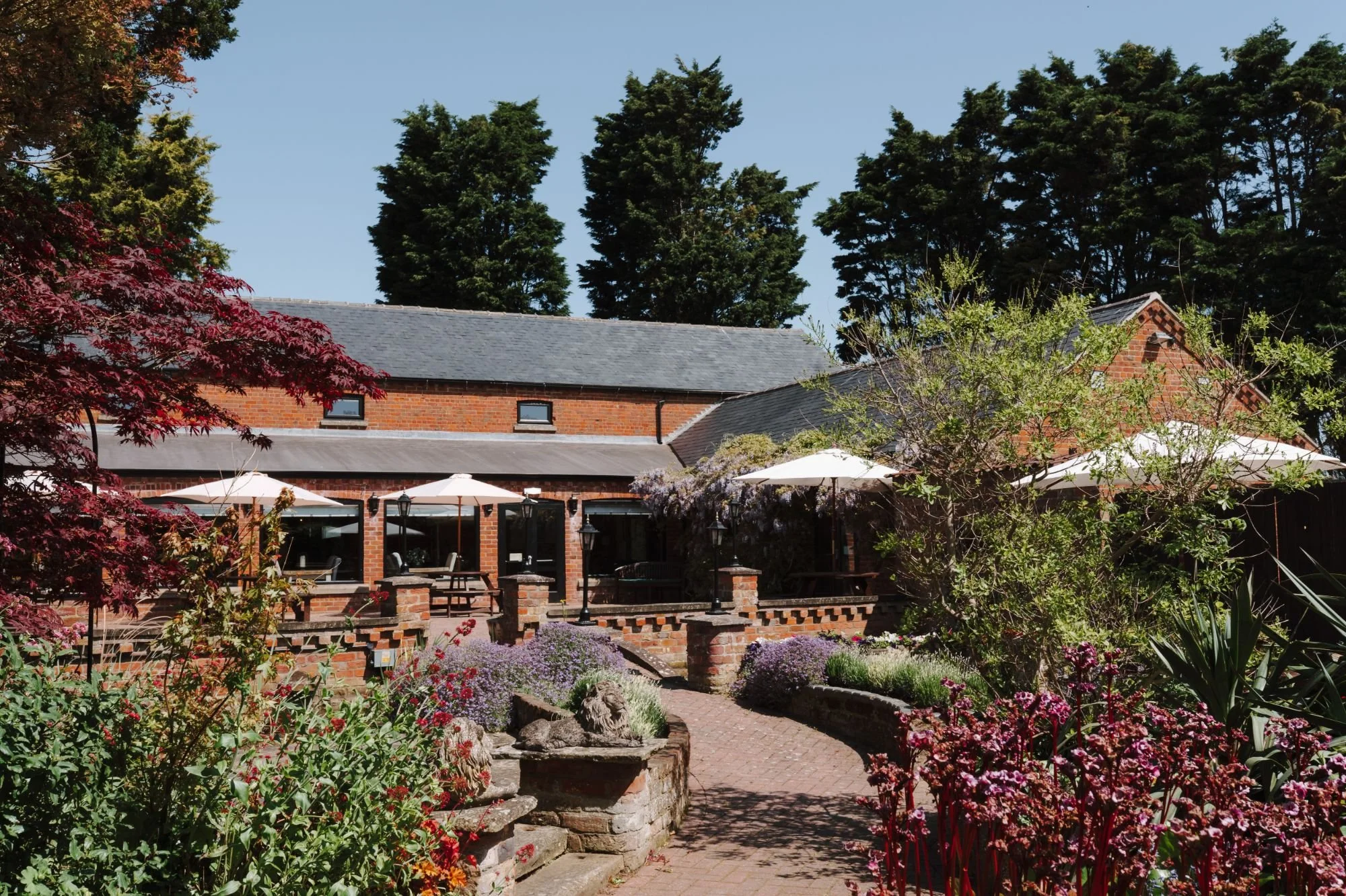 Whaplode Manor with outdoor seating, surrounded by colourful flowers, greenery, and tall trees under a clear blue sky.