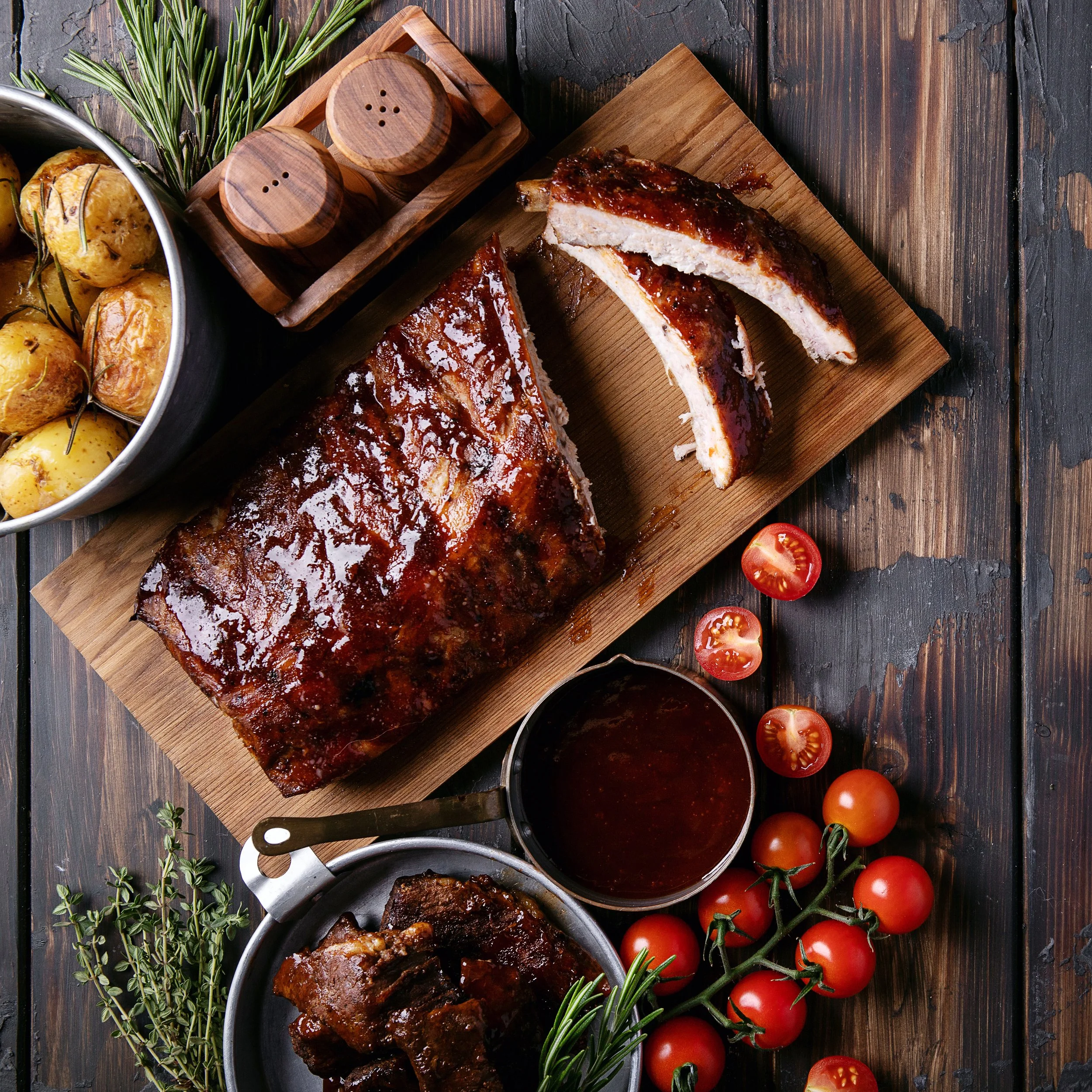 A rustic wooden table with a cutting board holding sliced barbecue ribs covered in sauce, a bowl of roasted potatoes with rosemary, cherry tomatoes still on the vine, fresh sprigs of rosemary and thyme, a small bowl of barbecue sauce, and wooden salt and pepper shakers.