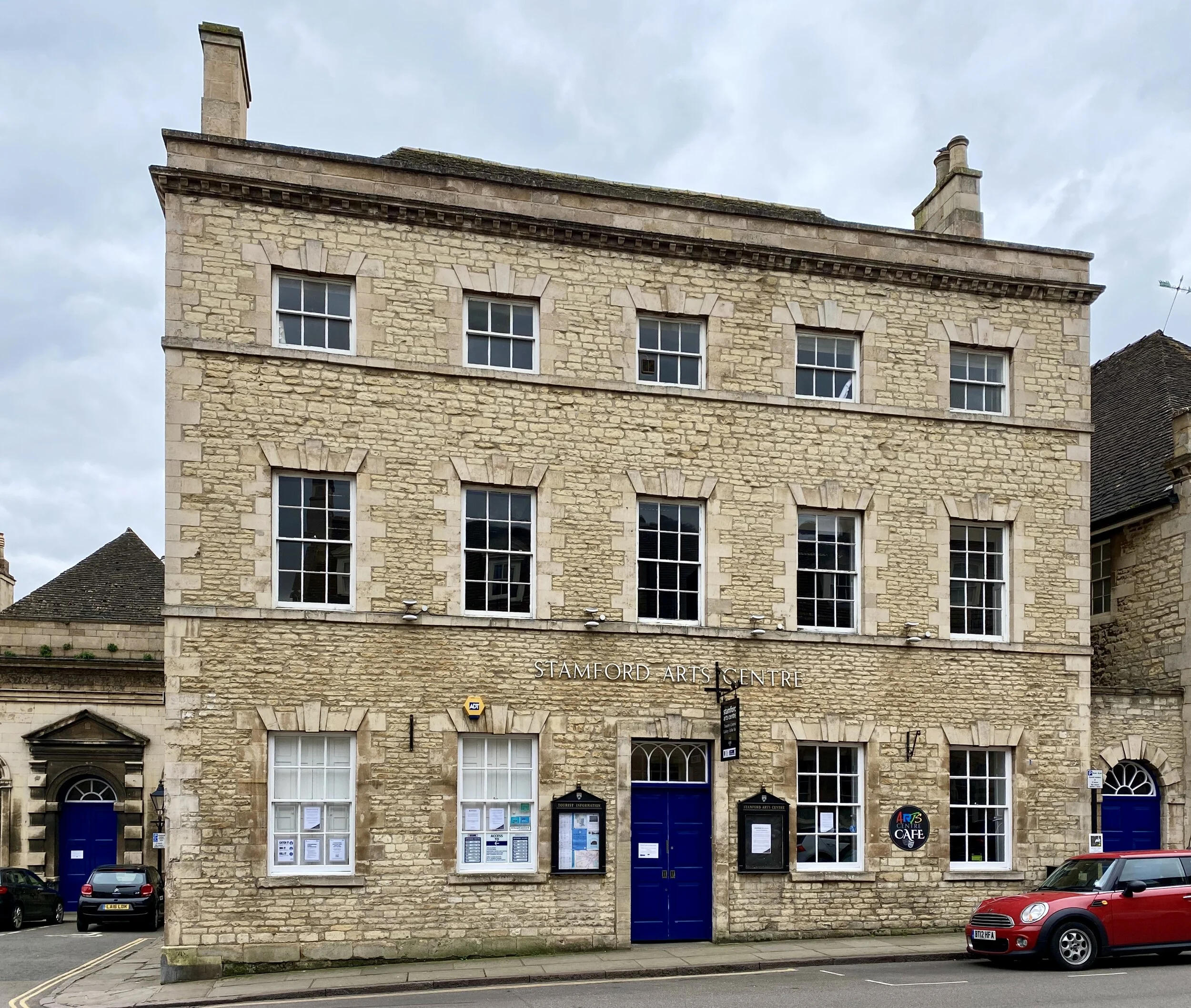 Stamford Arts Centre  with large windows and blue doors, signage for Stamford Arts Centre, and cars parked in front.