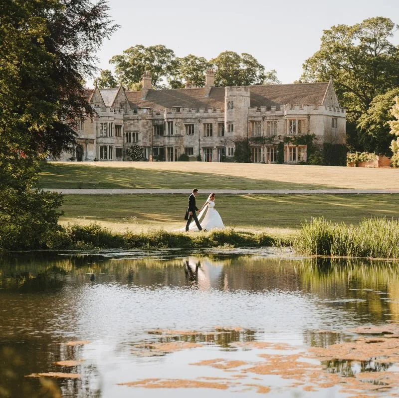 Irnhmam Hall with a bride and groom walking hand in hand on the grass near a pond, with a large stone mansion in the background surrounded by trees.