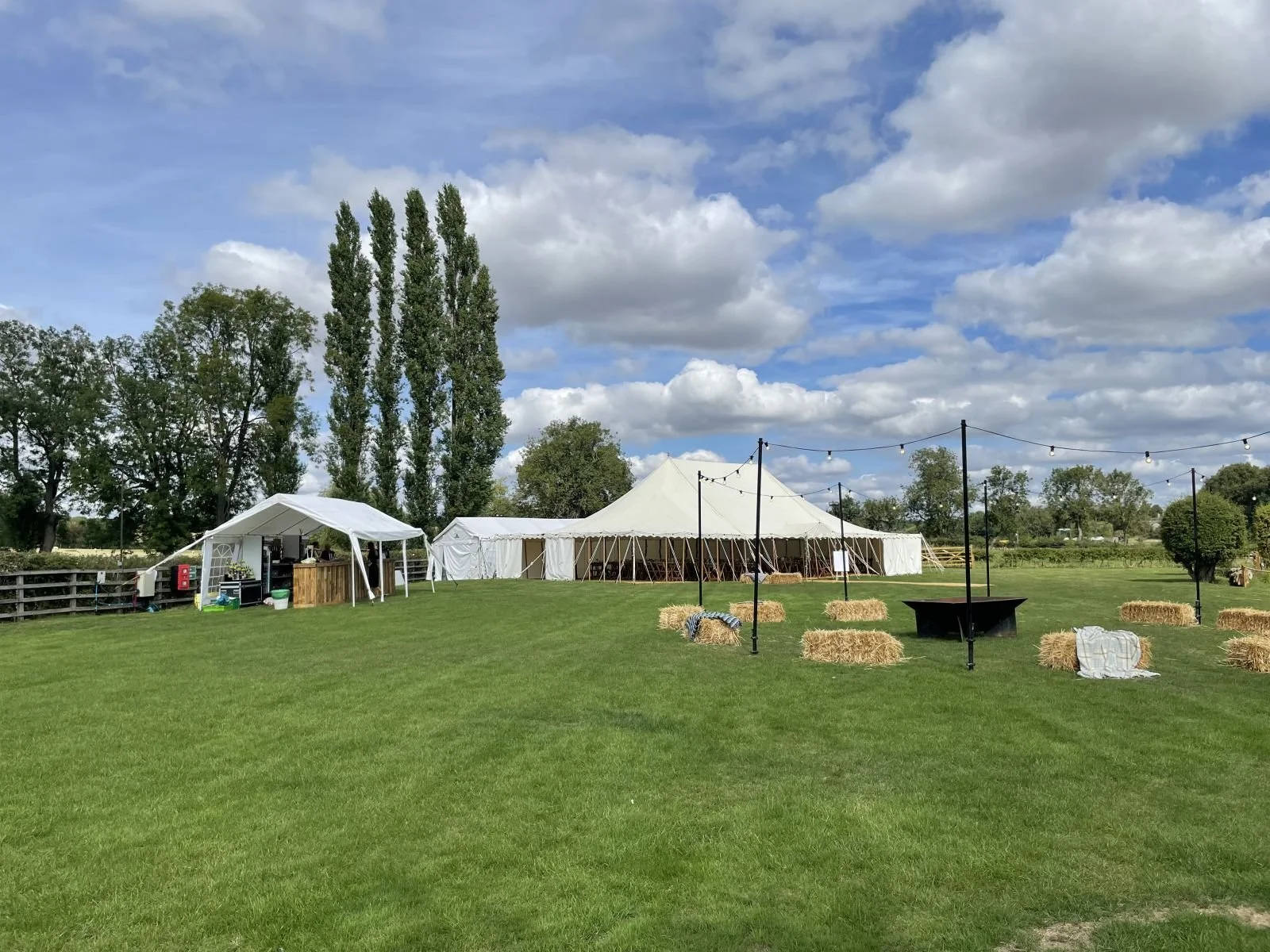 Berryfields Yarwell, with a large outdoor event setup with white tents, hay bales, string lights, and a green lawn under a partly cloudy sky.