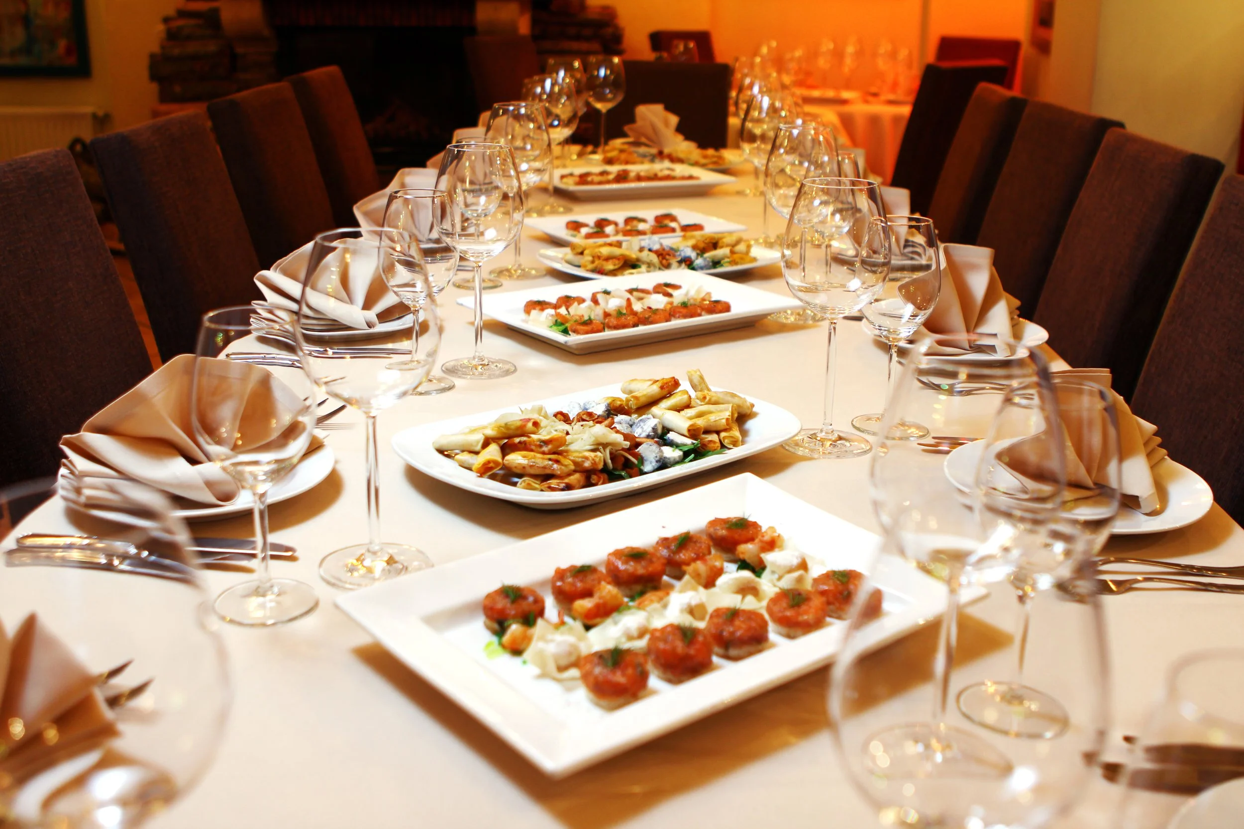 A long dining table set for a meal with plates of assorted appetizers, empty wine glasses, and folded napkins in a dimly lit restaurant or event space.