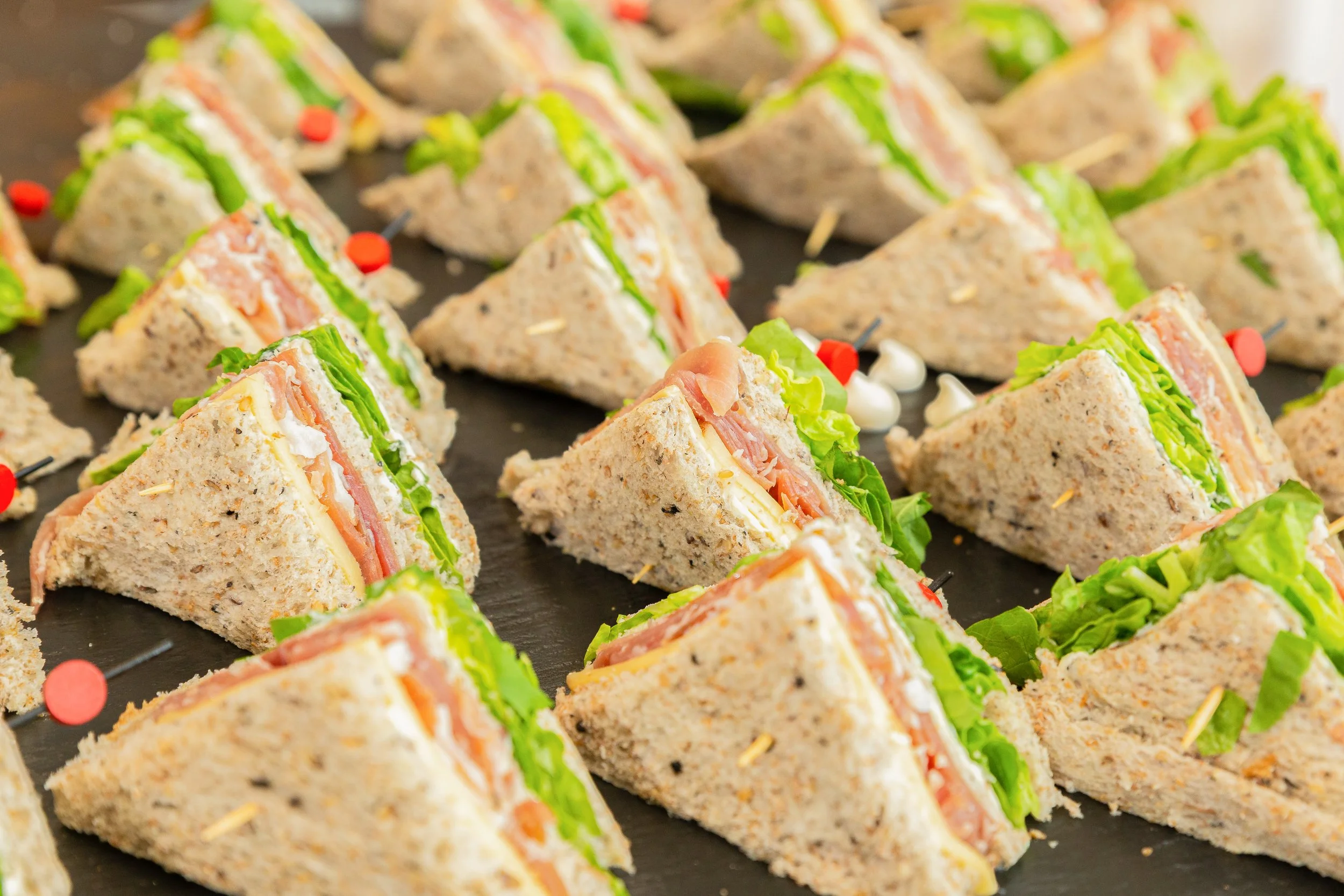 Assorted tea sandwiches with lettuce, tomato, cheese, and meat on sliced bread, arranged on a platter.