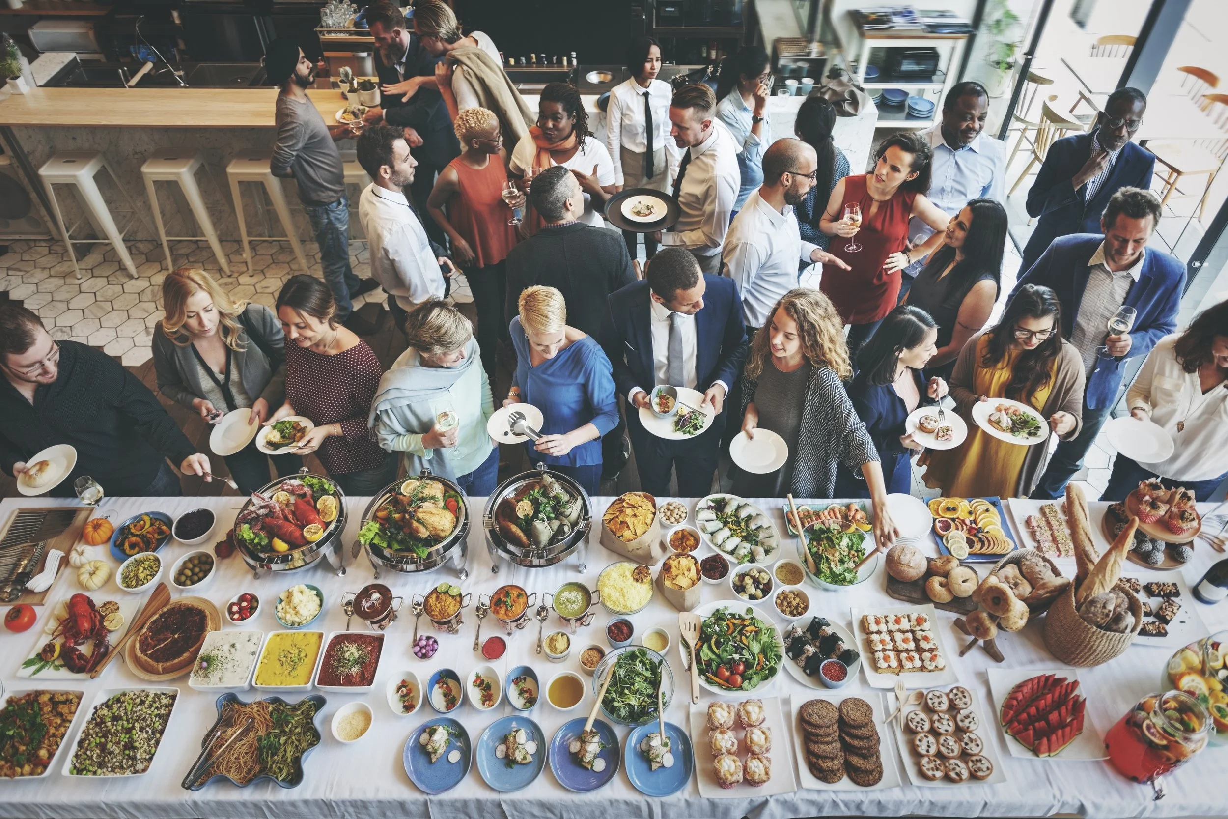 A large gathering of diverse people enjoying a buffet at a social event in a modern, well-lit restaurant or private dining area.
