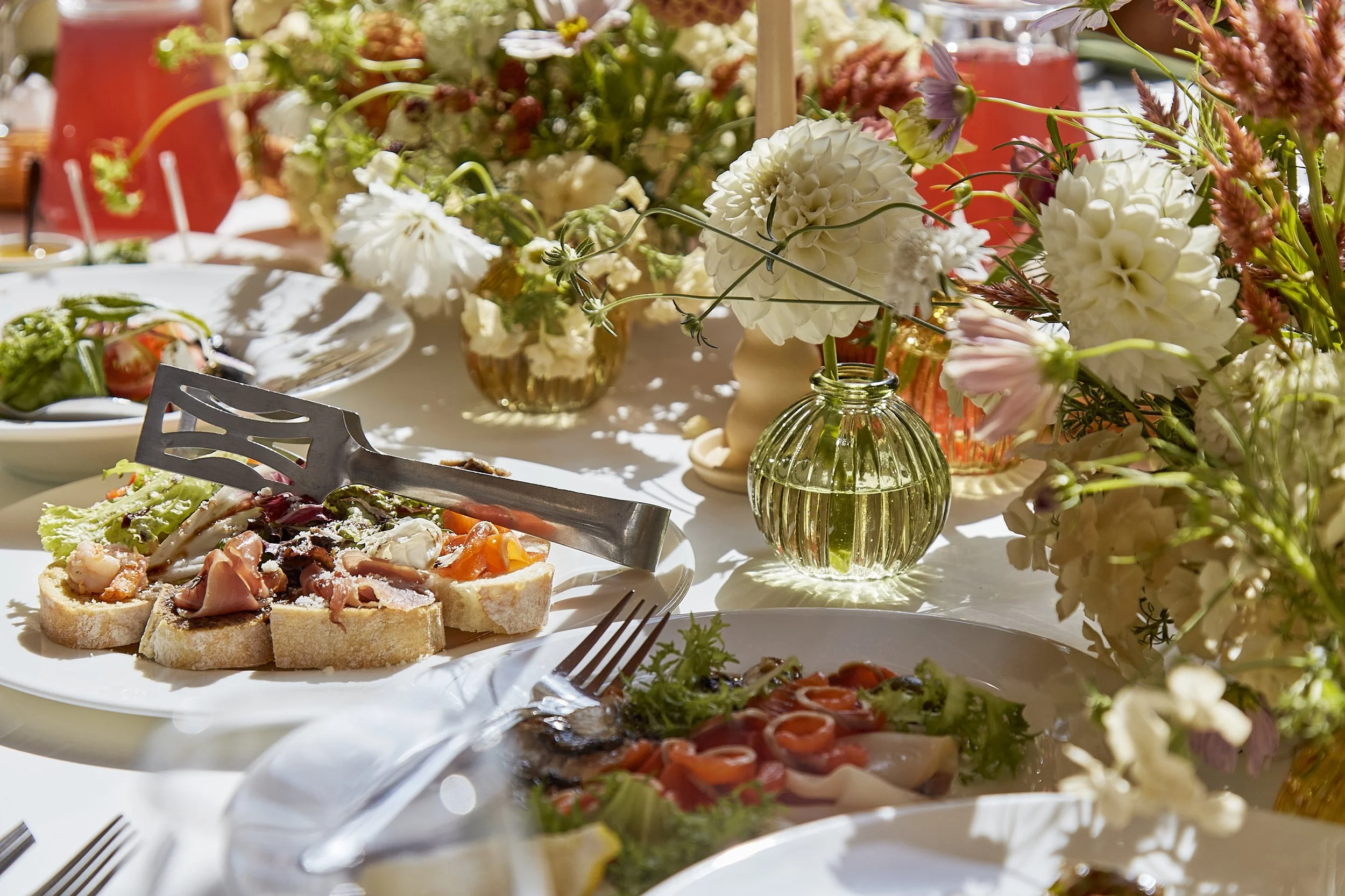 A table decorated with  a variety of dishes, including open-faced sandwiches with meat and vegetables, set for a meal outdoors in sunlight.