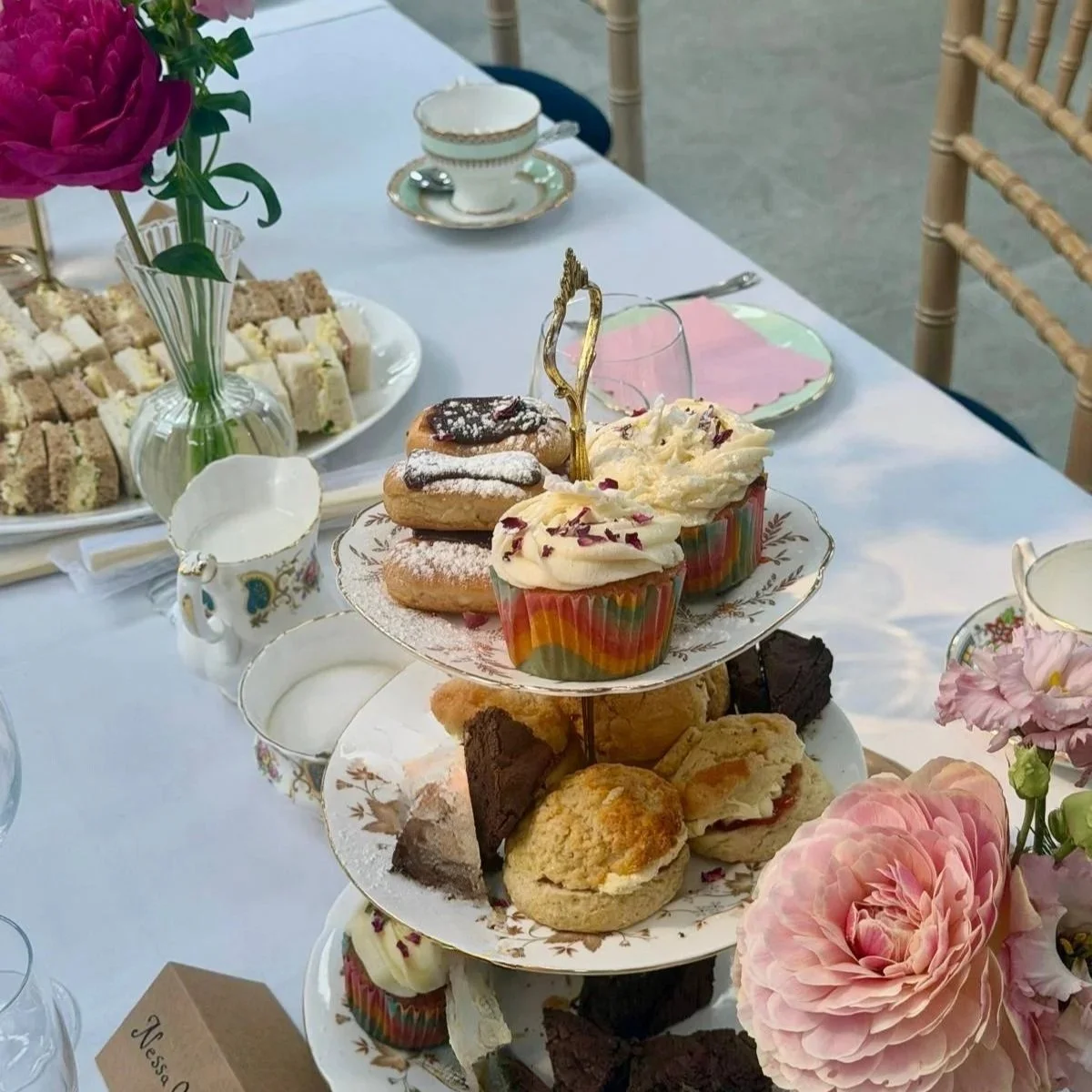 A tiered tray with assorted baked goods including cupcakes, cookies, and brownies, set on a table with floral decorations, teacups, and plates in a tea party setting.
