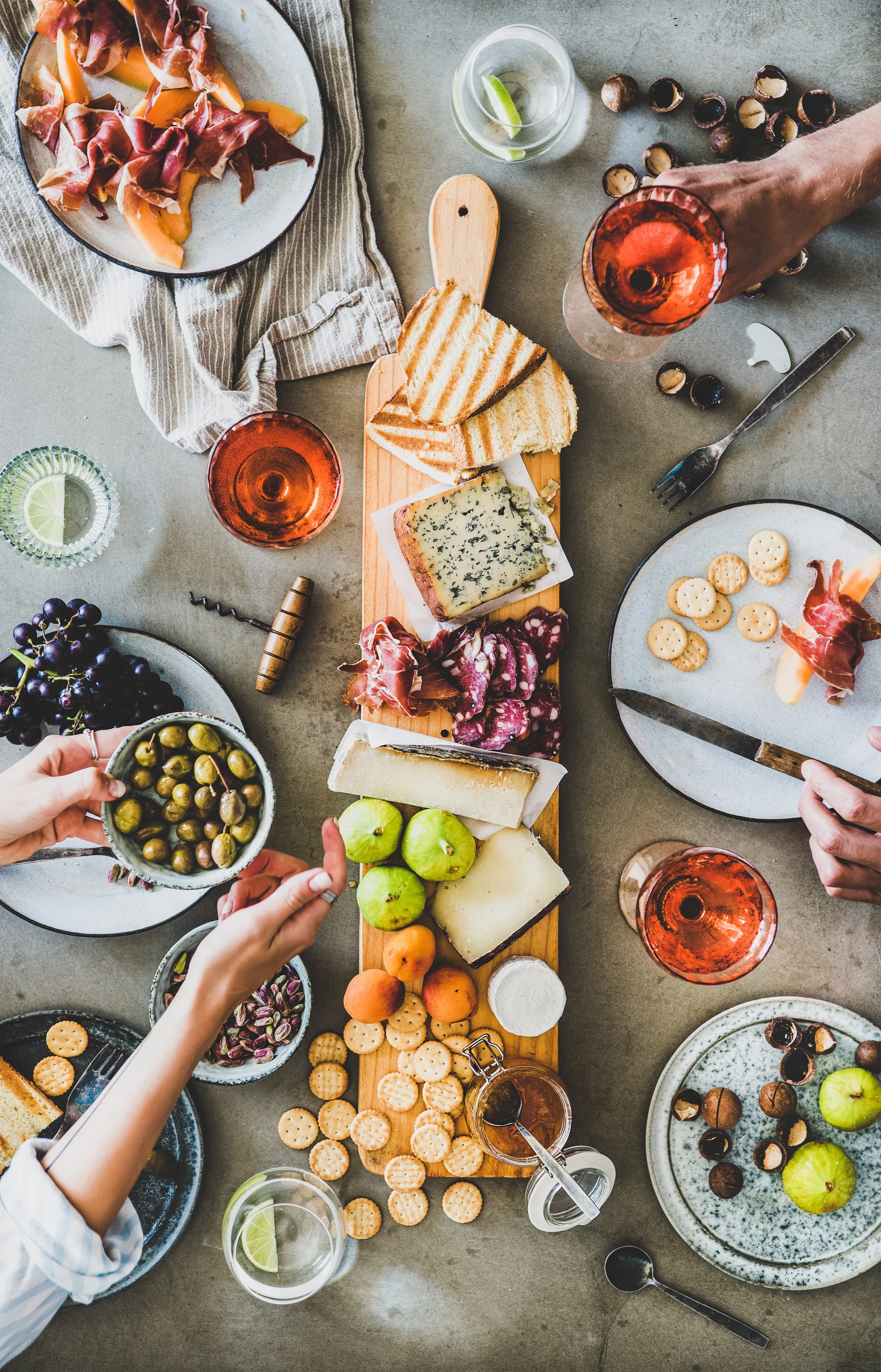 Overhead view of a table with assorted cheese, crackers, grapes, apples, and glasses of water and rosé wine. Several hands are reaching for food.