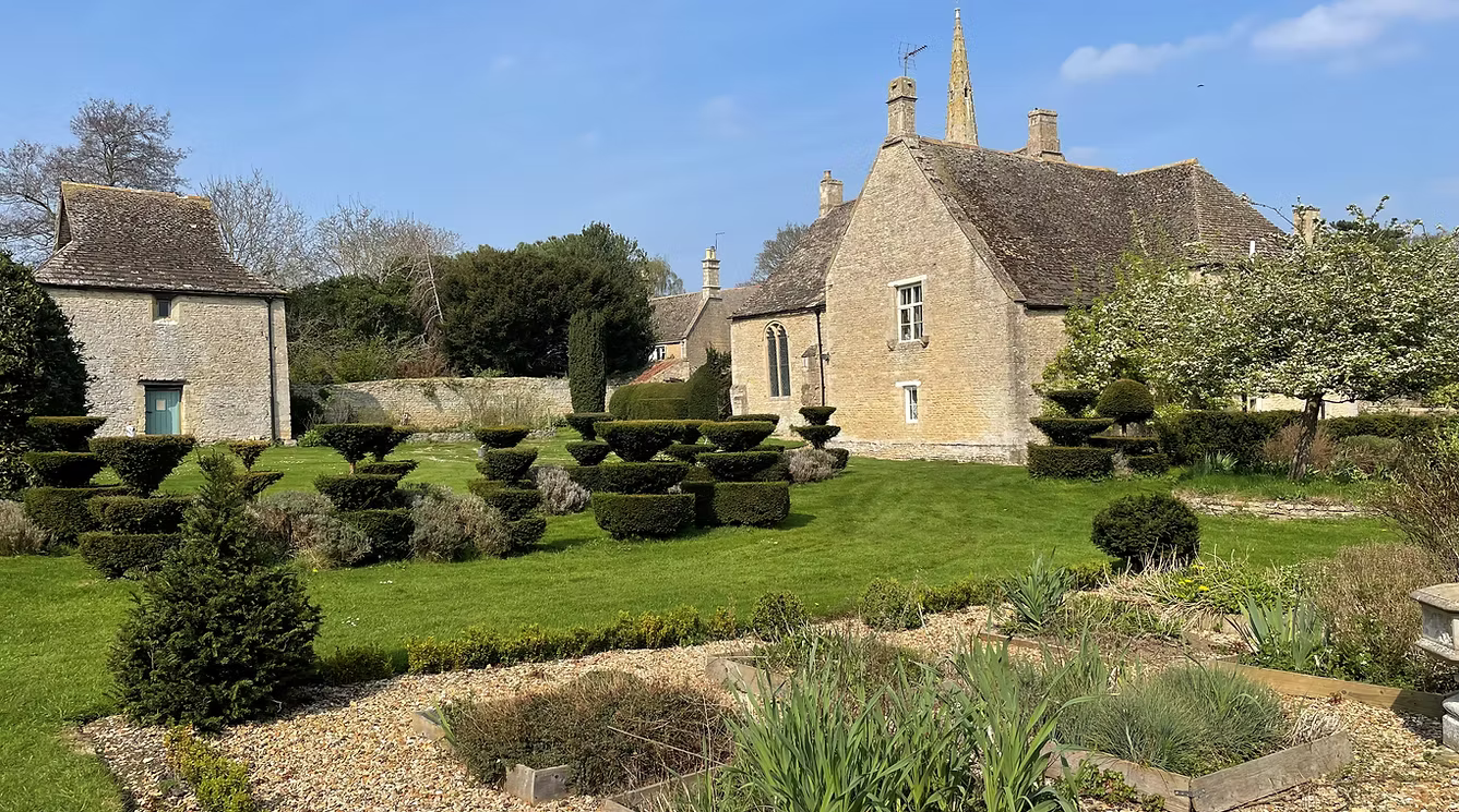 Prebendal Manor with a picturesque garden with trimmed bushes, a green lawn, and old stone buildings in the background on a sunny day.