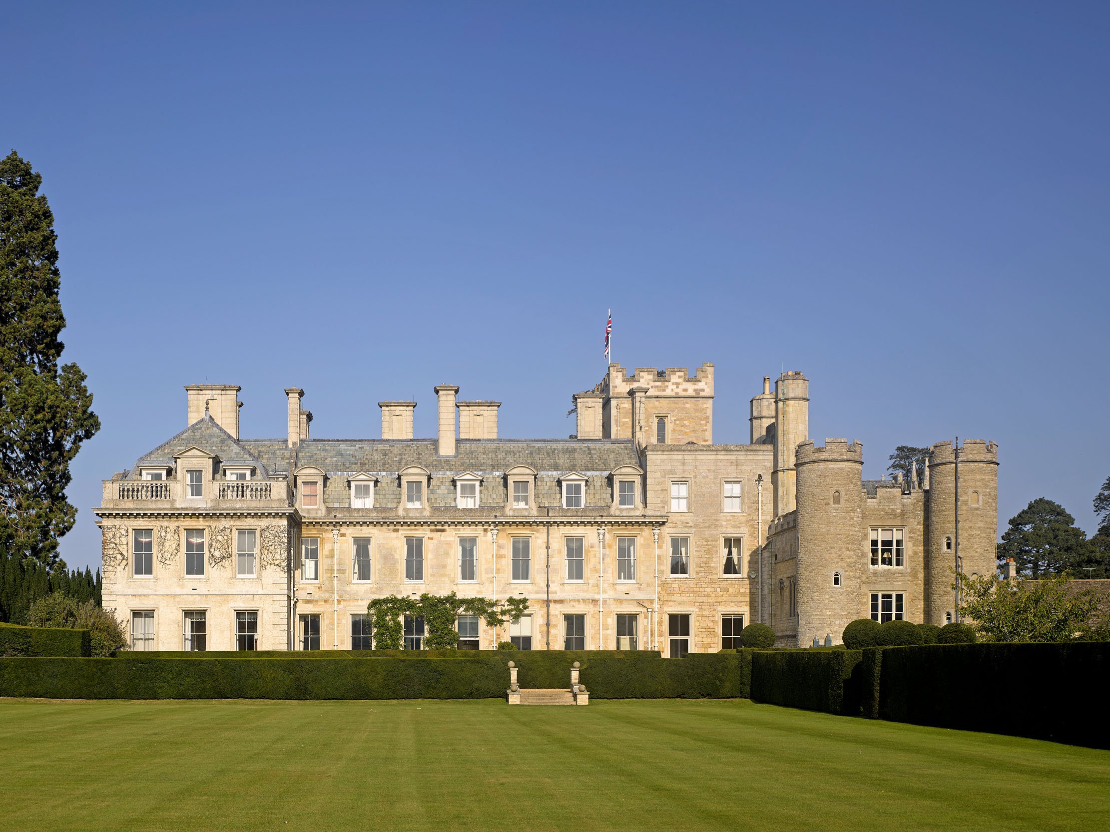 Elton Hall with multiple turrets and a flag on top, surrounded by a well-maintained lawn and trees, under a clear blue sky.