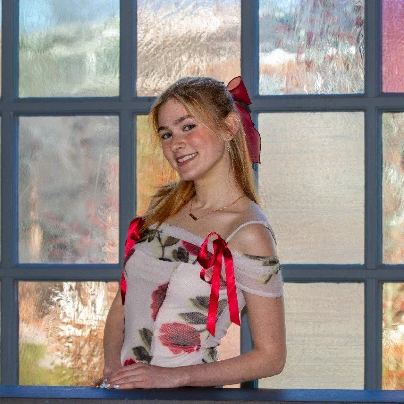 Smiling young woman with a red hair bow wearing a floral off-shoulder top in front of frosted glass windows