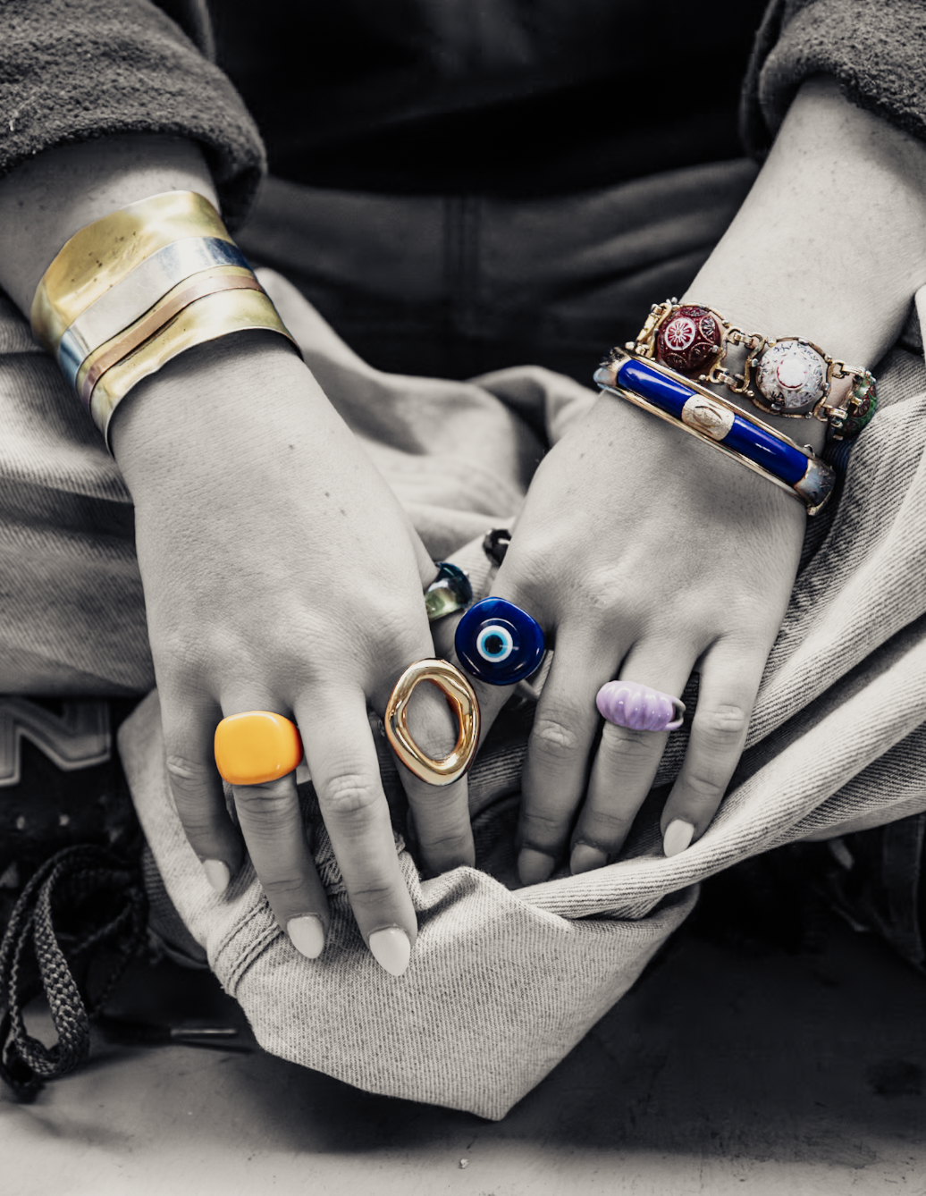 Close-up of hands wearing colorful statement rings, stacked bracelets, and cuff jewelry over a canvas bag