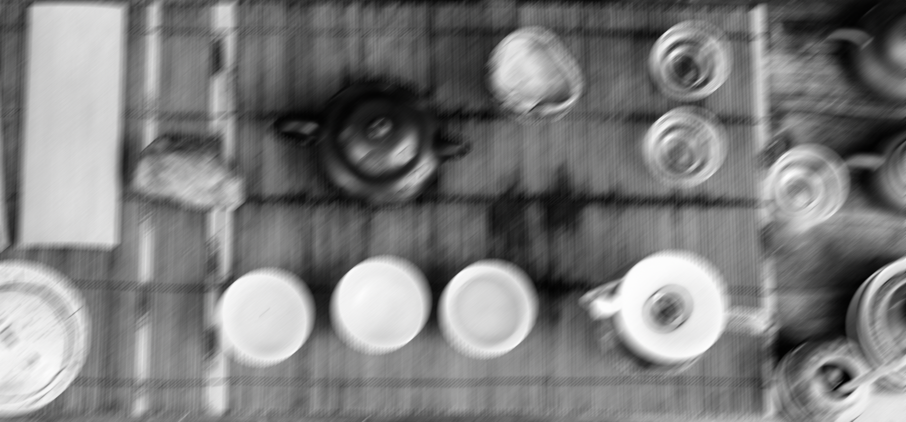 Blurred black-and-white overhead view of assorted dishes, bowls, cups, and kitchenware on a table