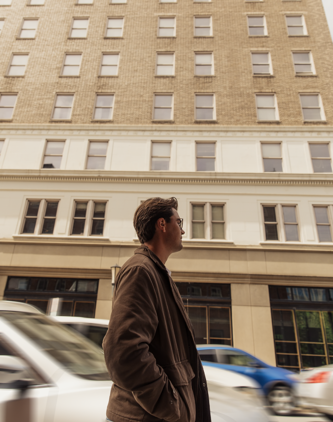 Man wearing glasses and a brown jacket walking past a city building with blurred traffic.