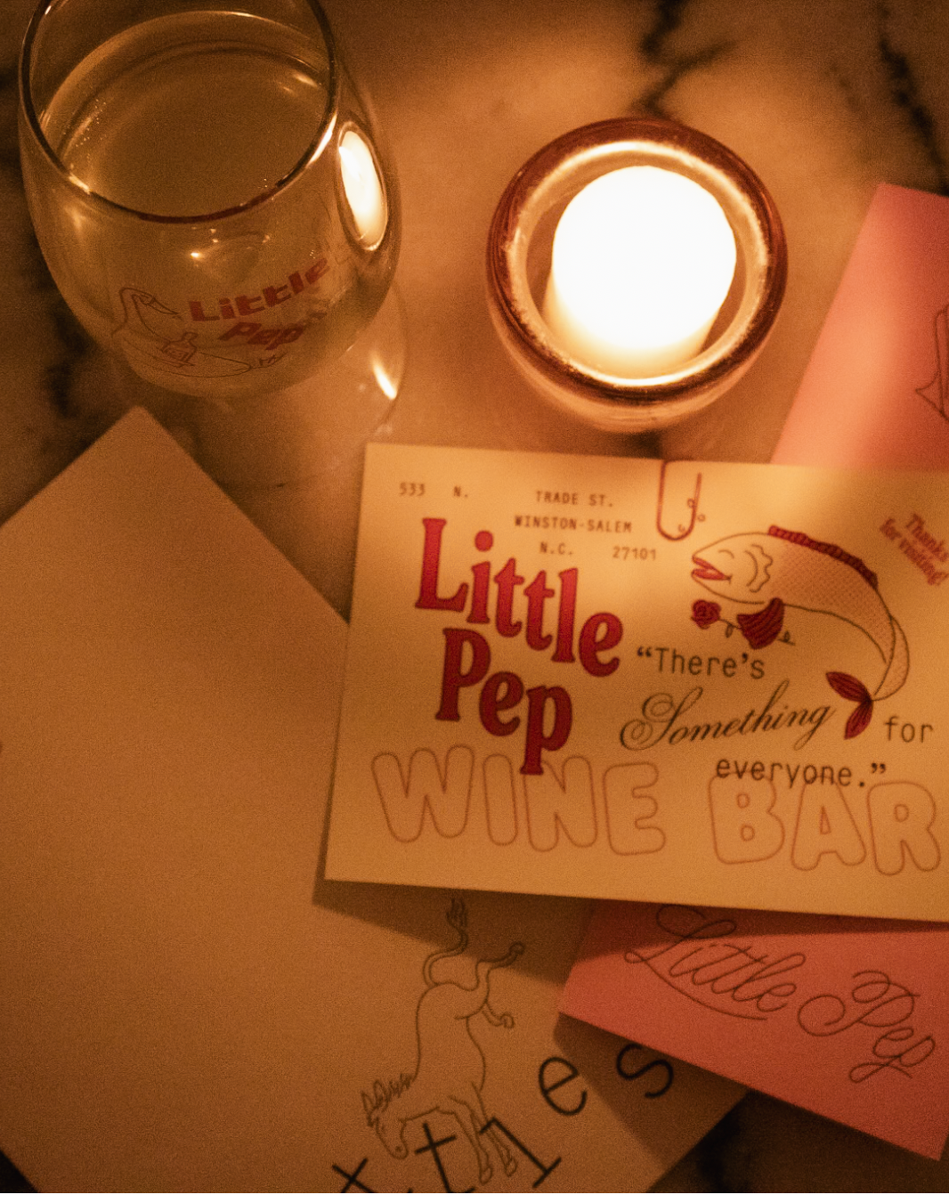 Warmly lit tabletop with a glowing candle, wine glass, and vintage Little Pep Wine Bar menu cards.