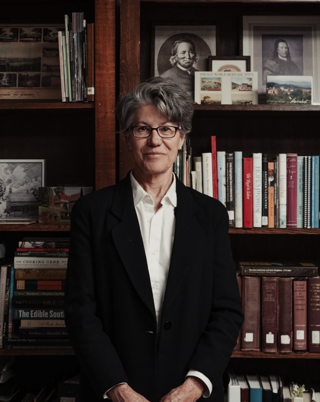 Portrait of a smiling woman in glasses and a black blazer standing in front of a bookshelf