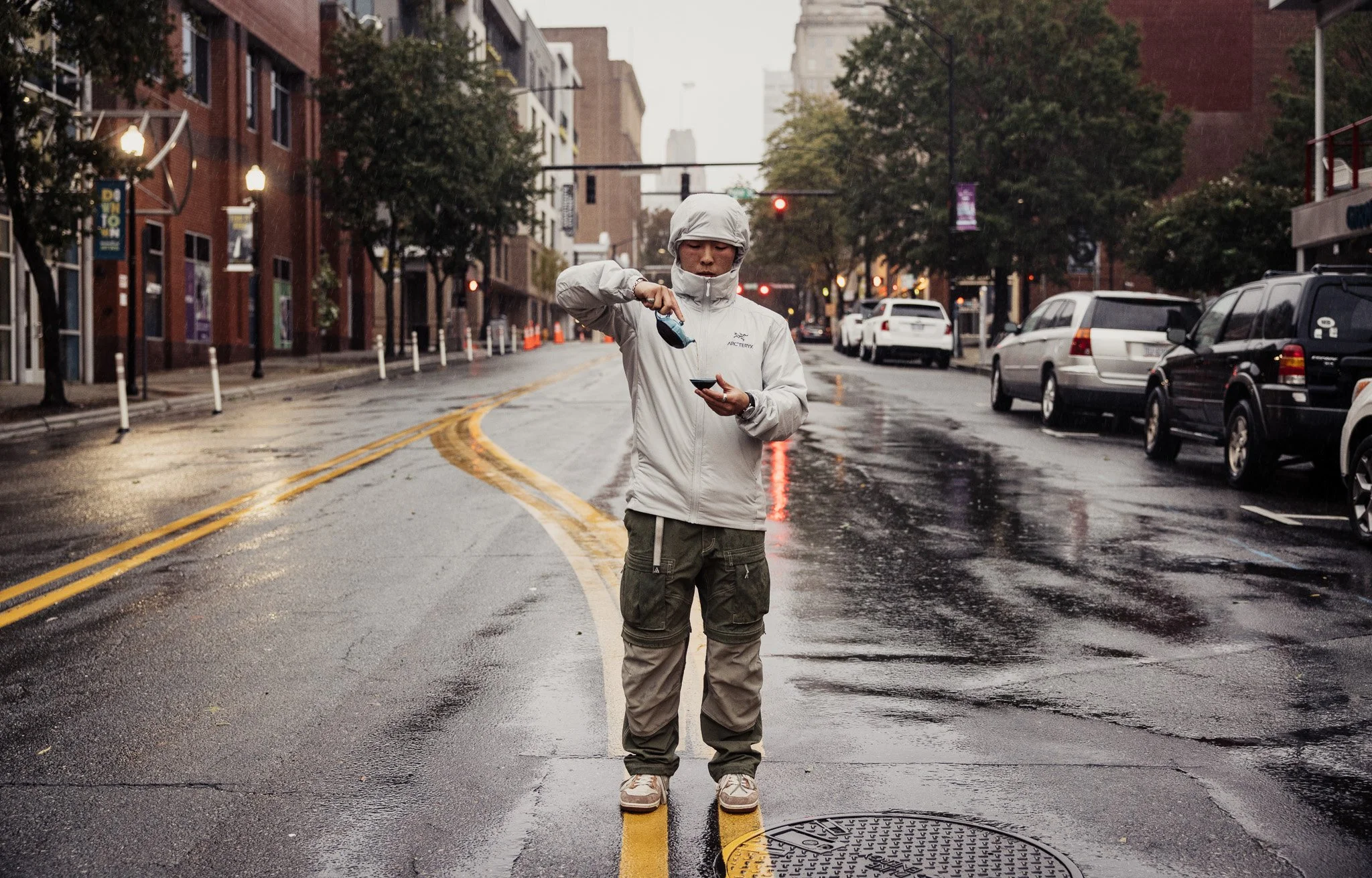 Person in a white hooded jacket standing on double yellow lines in a rainy city street, holding a smartphone and a small object.