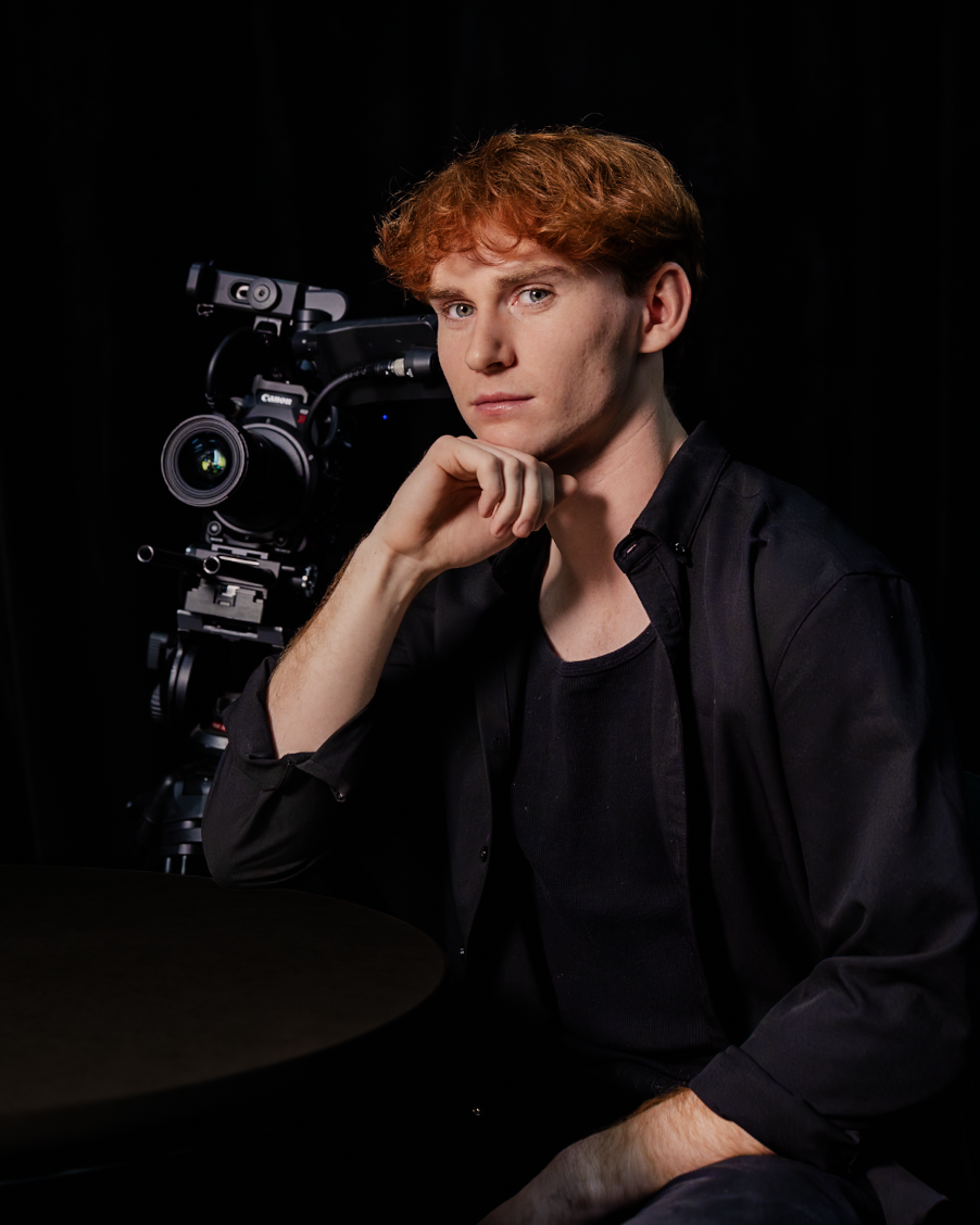 Red-haired young man posing beside a professional video camera against a black background