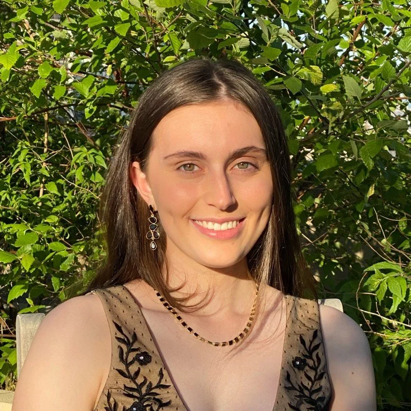Smiling woman with long brown hair wearing a sheer embroidered dress and jewelry outdoors among leafy greenery