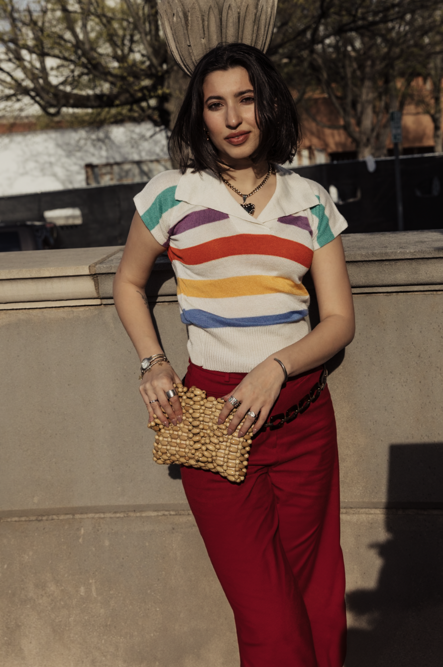 Woman posing outdoors in a colorful striped top, red pants, and a wooden-bead handbag