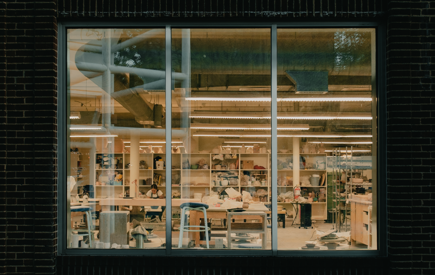 Pottery studio viewed through a large window, with worktables, shelves of ceramics, tools, and bright fluorescent lights