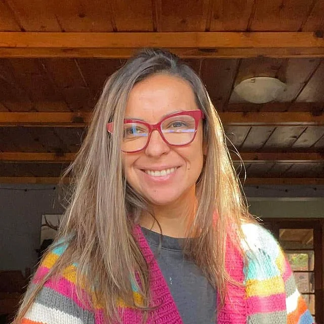 Woman with long blonde hair, wearing red glasses, a gray shirt, and a colorful striped cardigan, smiling indoors with wooden ceiling and window in the background.