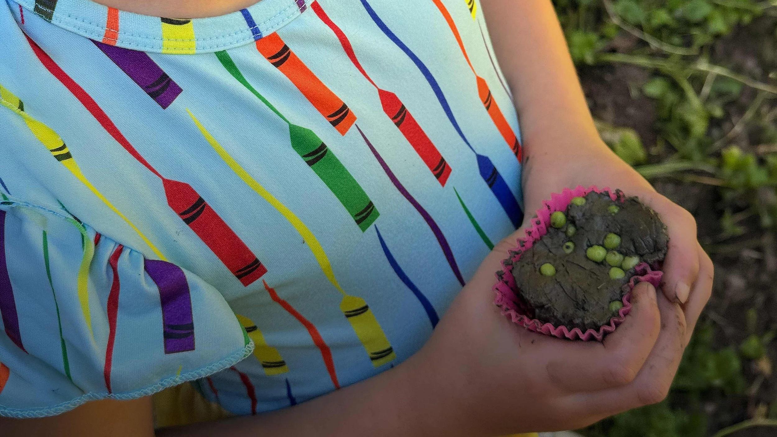 A child's arm holding a small roo cube toy covered in green and pink beads, while wearing a sleeveless shirt with colorful crayon patterns.