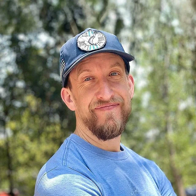 A smiling man with a goatee and mustache wearing a blue baseball cap and a blue t-shirt outdoors with trees in the background.