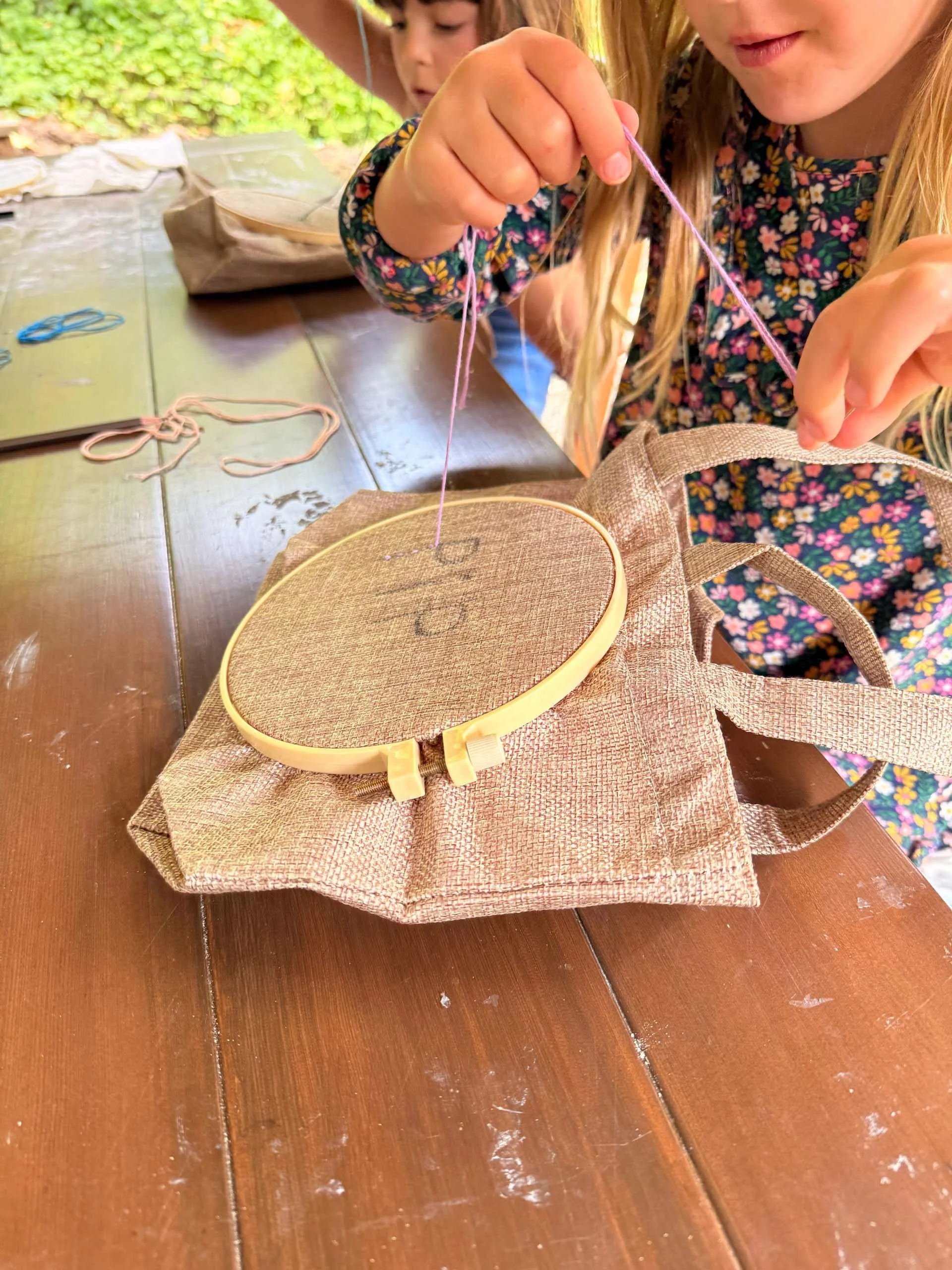 Two children, a girl with long blonde hair and a boy with short brown hair, are sitting at a wooden table. The girl is holding a piece of embroidery thread and appears to be working on a craft project, with an embroidery hoop on the table in front of her. Various pieces of thread are on the table, and there is a bag nearby. The background shows outdoor greenery.