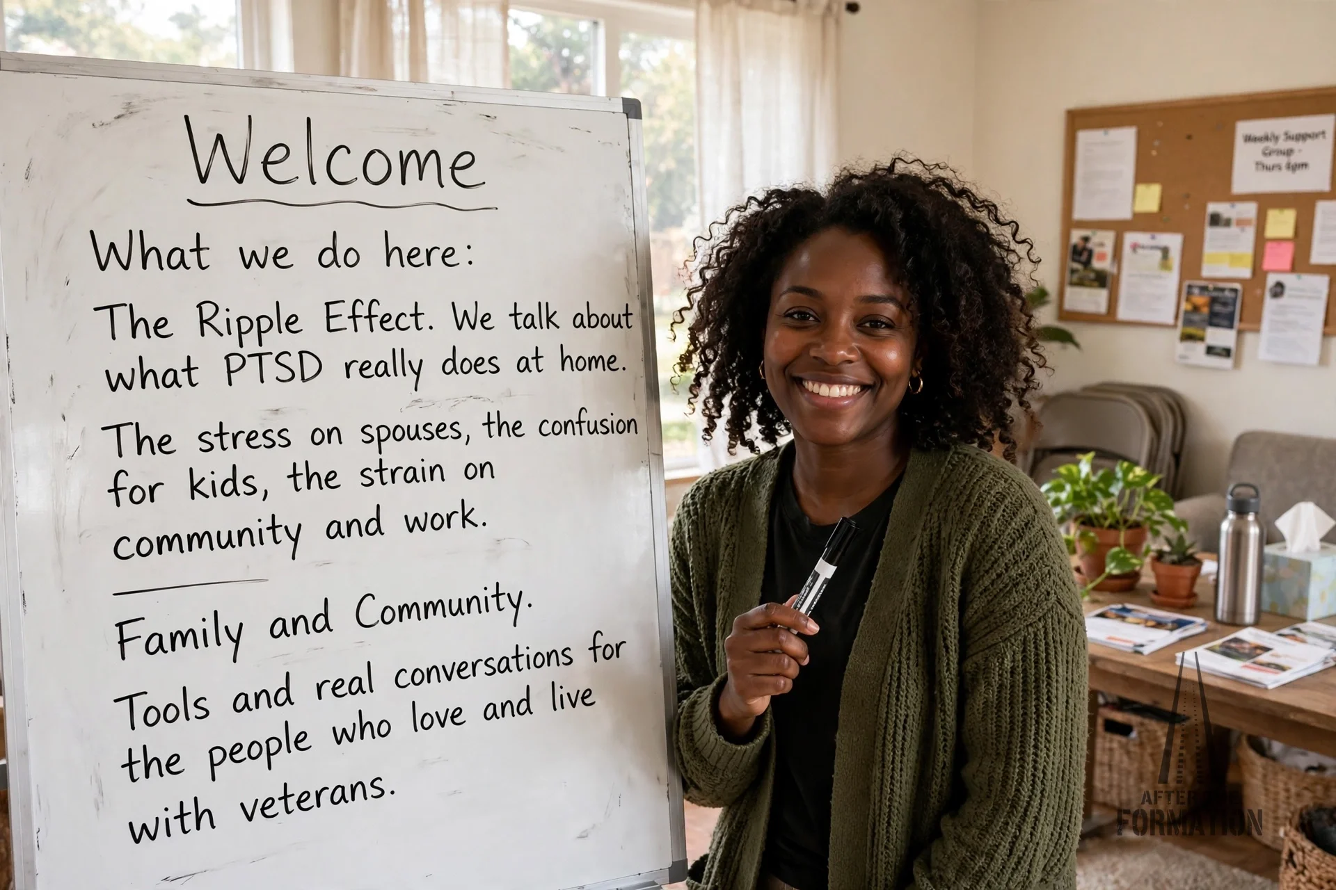 A woman with dark curly hair and earrings, smiling, standing next to a whiteboard in a well-lit room with large windows and bulletin boards. She holds a black marker in her hand.