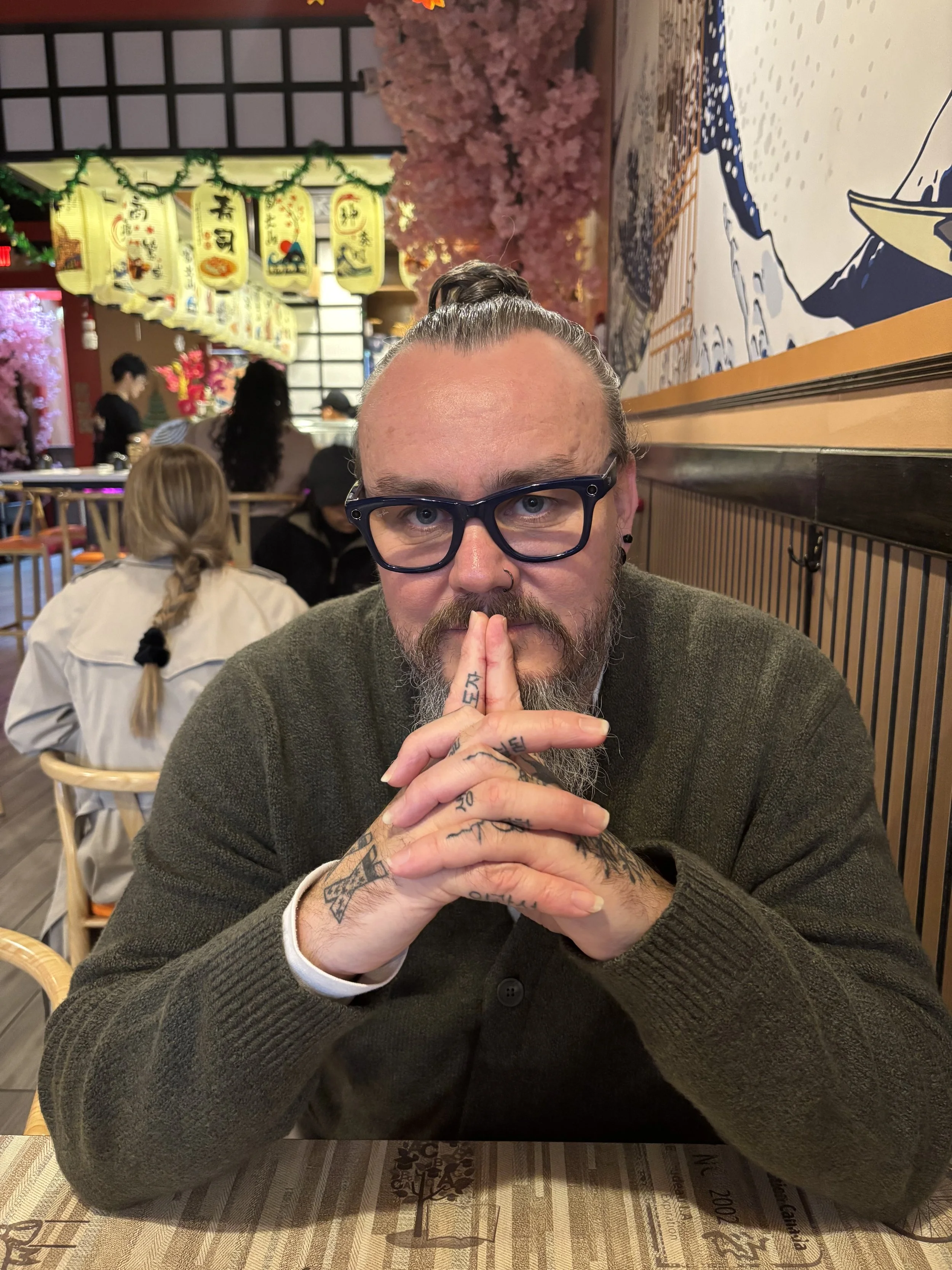 A man wearing glasses and with tattoos on his hands is sitting at a restaurant table with his hands pressed together near his mouth, looking into the camera. The restaurant has Asian-themed decor with lanterns hanging from the ceiling, pink cherry blossom trees, and a mural of mountains and ocean.