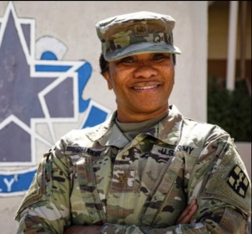 A smiling African American female soldier in camouflage uniform with arms crossed, standing outside near a wall with a blue and white emblem or logo.