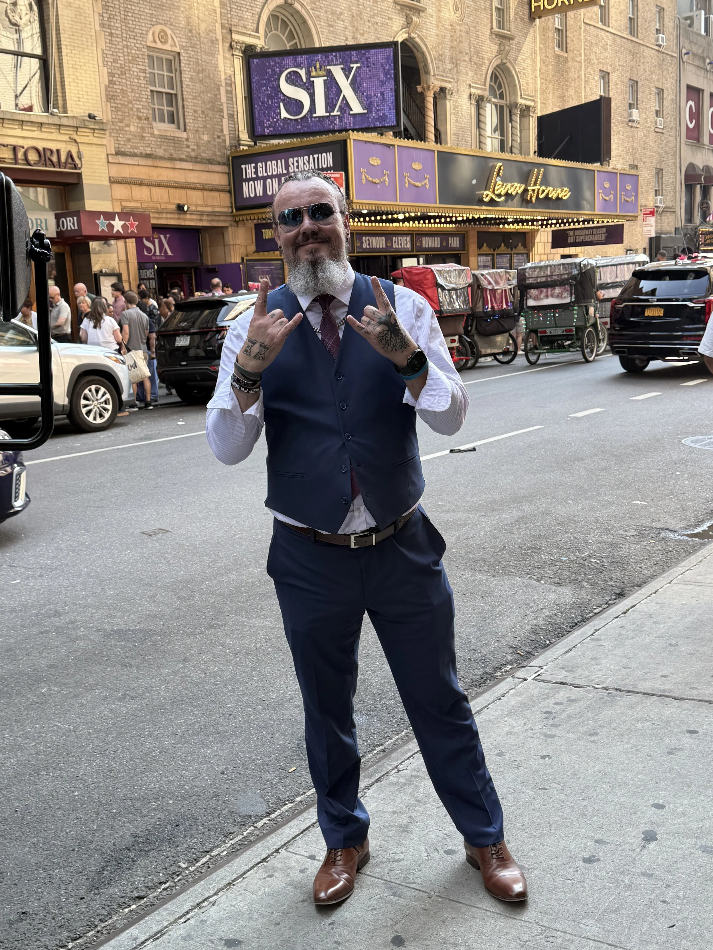 A man with sunglasses and a beard stands on a city sidewalk, making a hand gesture with both hands. He is dressed in a blue vest, white shirt, red tie, and brown shoes. There are cars and pedestrians in the background, with a theater marquee advertising 'Six' and various signs.