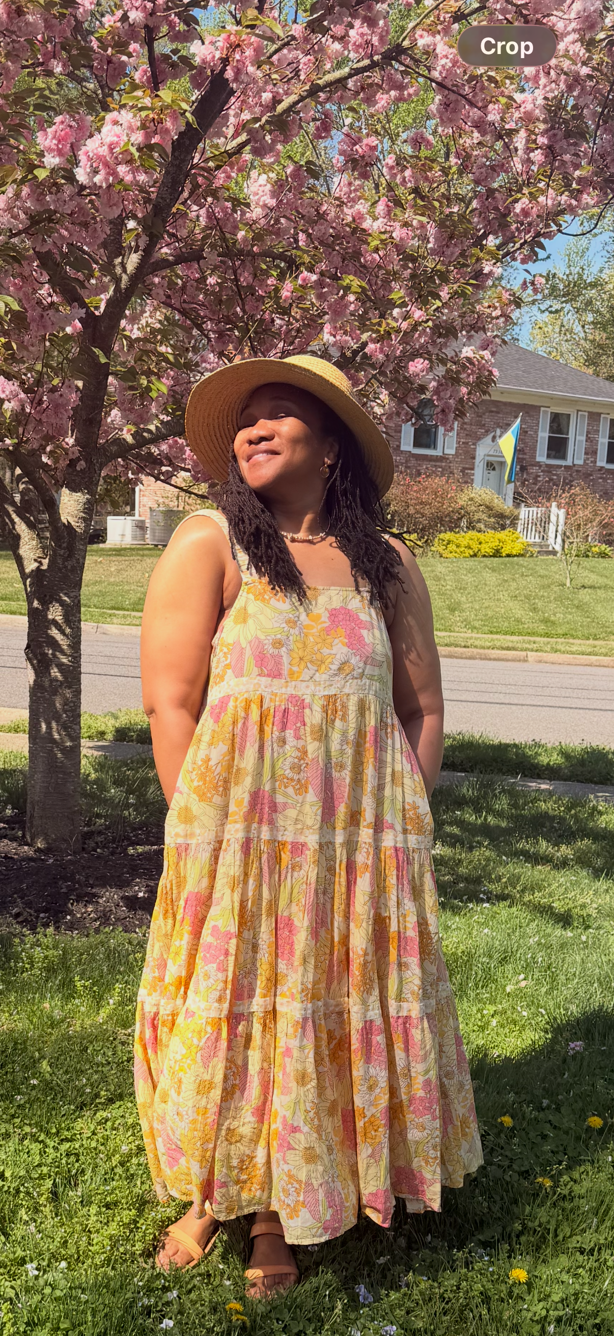 A woman in a yellow floral dress and wide-brimmed straw hat stands outdoors on a sunny day, with pink blossoms on a tree behind her and a house in the background.