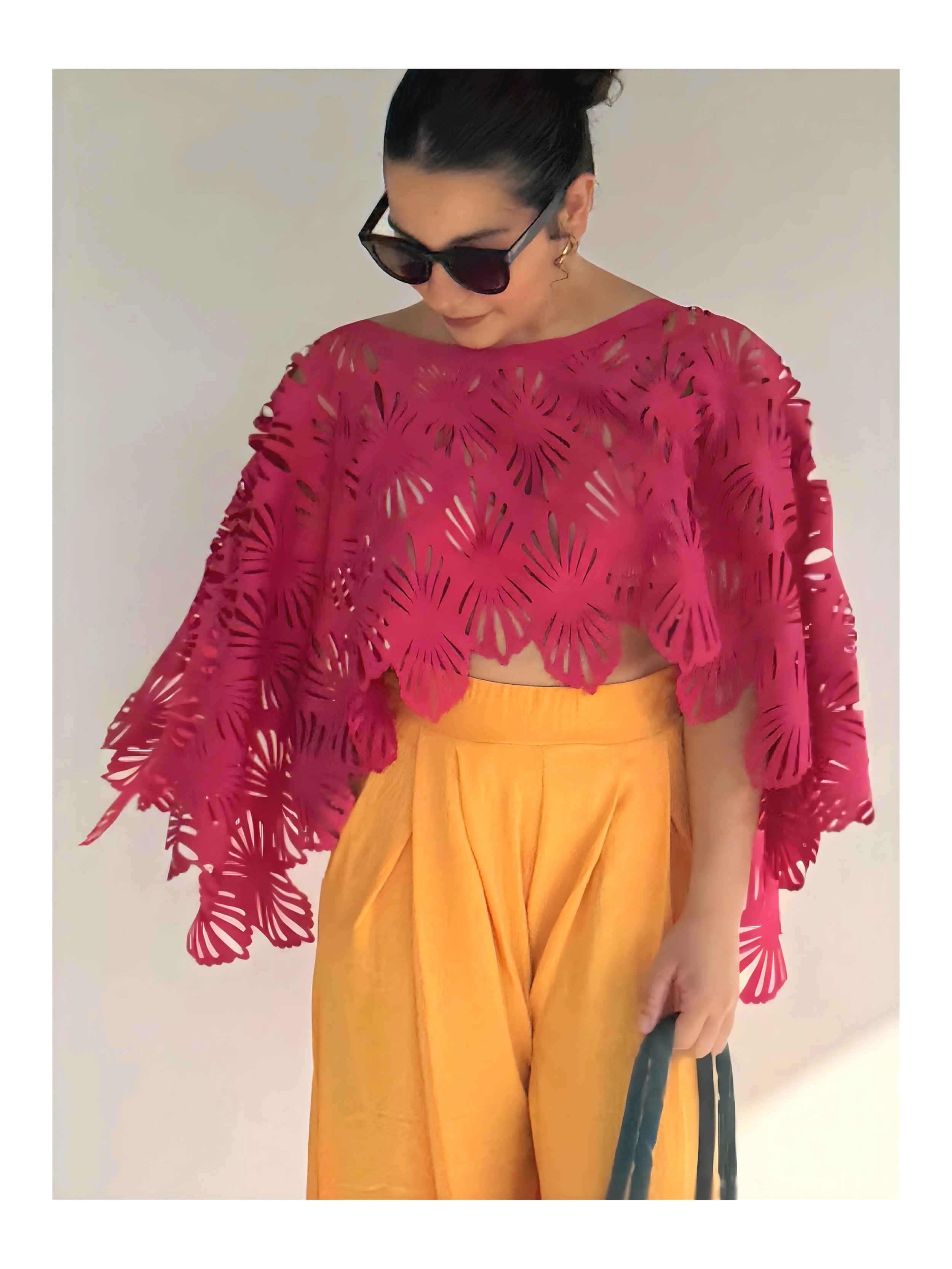 A woman wearing sunglasses, a pink laser-cut top, and mustard yellow high-waisted pants, holding a dark handbag.
