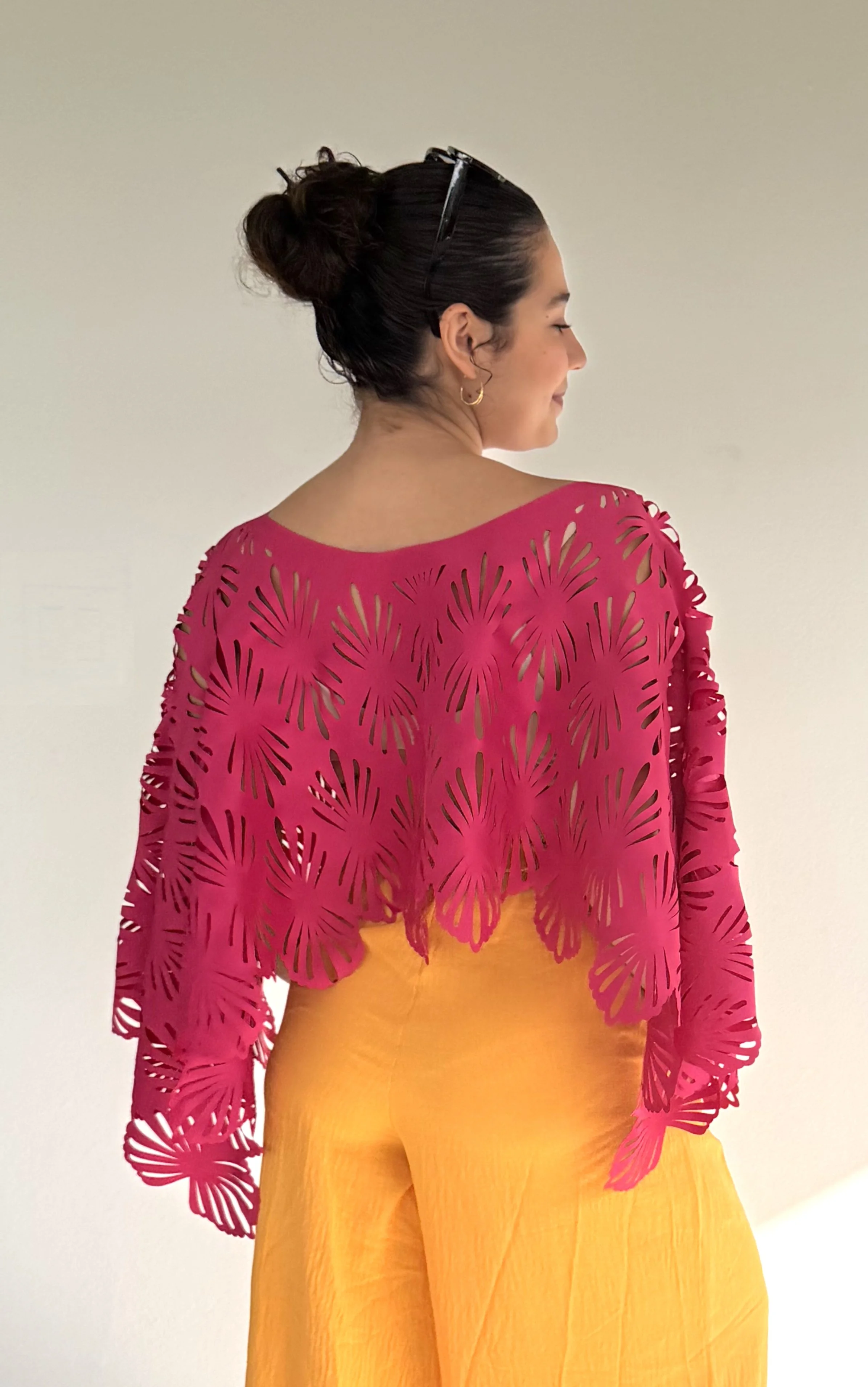 Woman from behind with dark hair in bun, wearing pink cut-out jacket and yellow pants, standing against a plain white background.