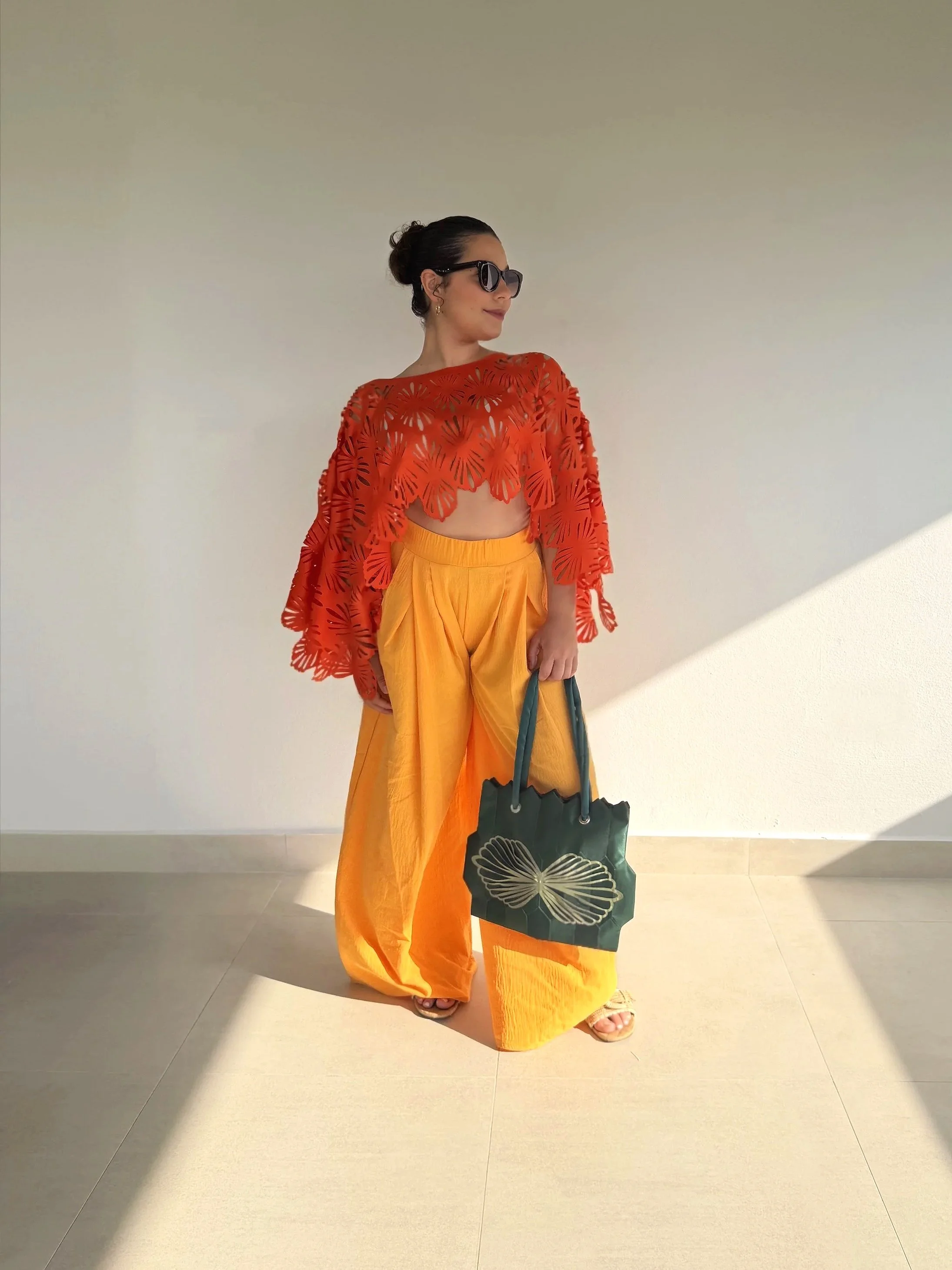 A woman wearing sunglasses, a red floral top, and yellow wide-leg pants, holding a dark green bag with a butterfly design.