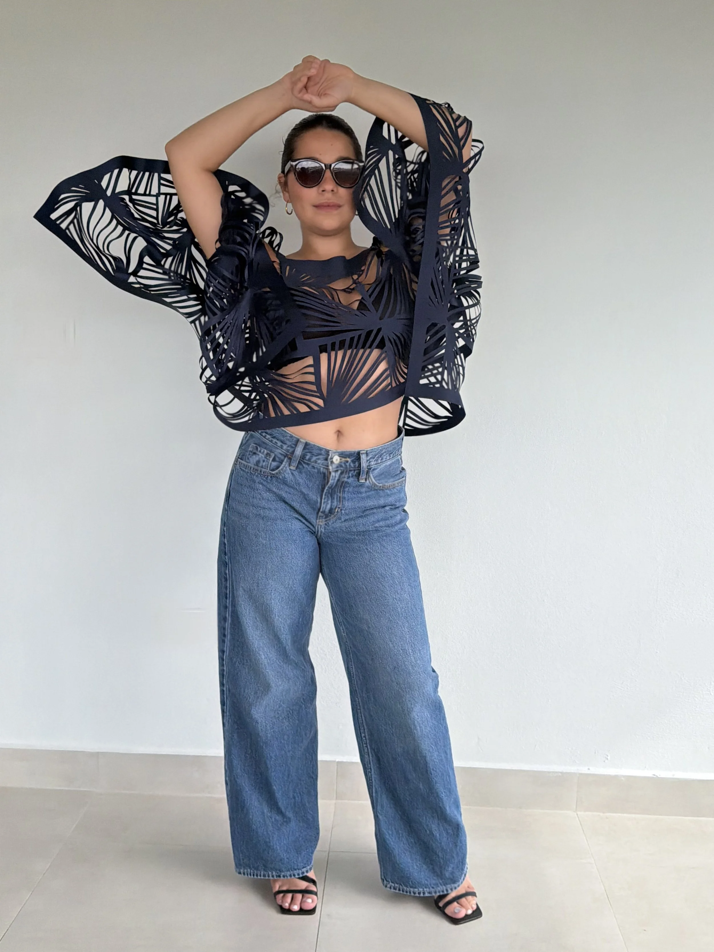 Woman wearing black sunglasses, a black cut-out top with a geometric pattern, blue jeans, and black sandals, standing against a plain white wall.