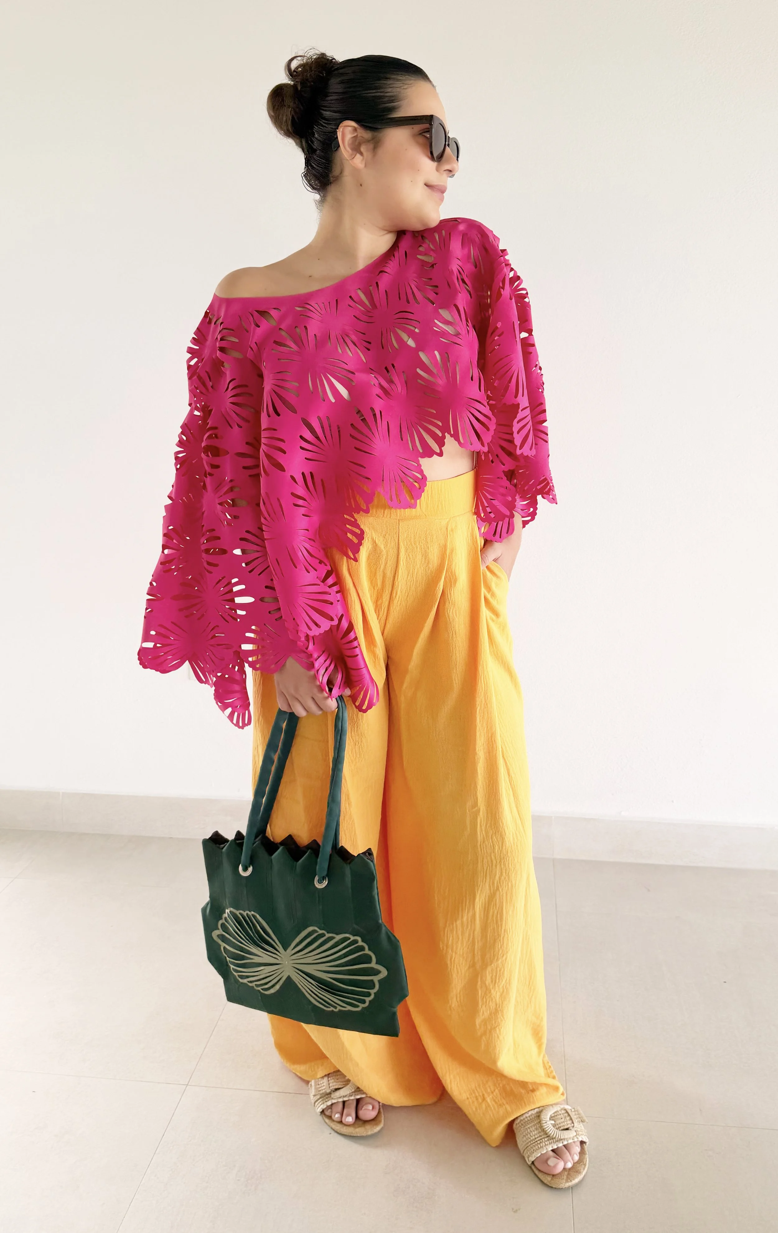 A woman with dark hair tied up, wearing glasses, a bright pink off-the-shoulder top with leaf cutouts, yellow wide-leg pants, beige shoes, and holding a green bag with a butterfly design, standing against a plain white wall.