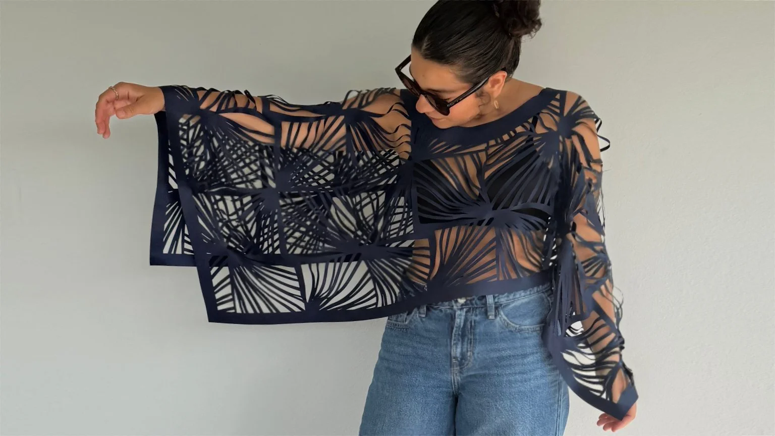 Woman wearing a navy blue, laser-cut, patterned top with long sleeves, paired with blue jeans, standing against a plain wall, adjusting or showing the design of her top, while wearing sunglasses and earrings.