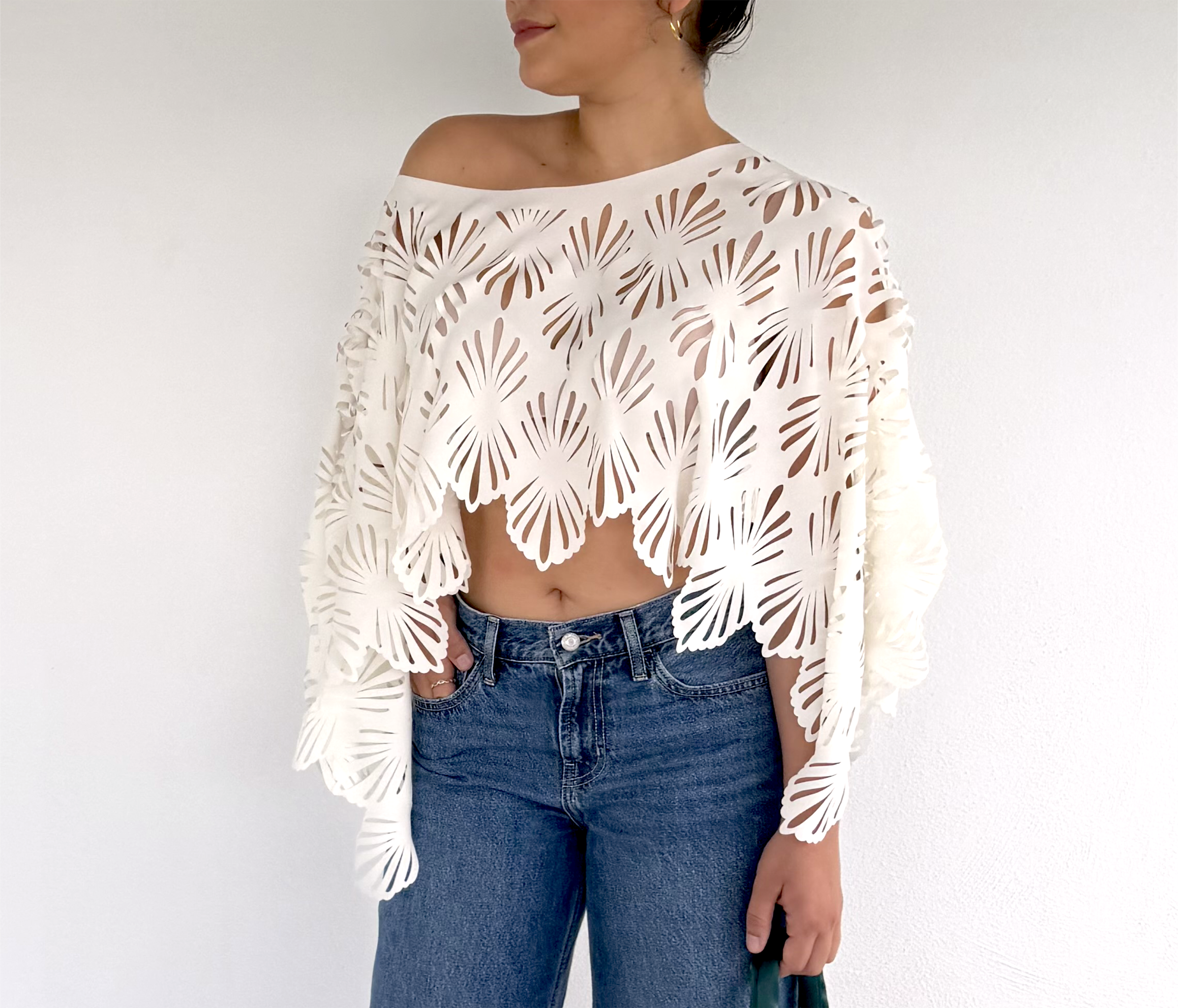 Woman wearing a white off-the-shoulder top with a laser-cut floral pattern and blue jeans, standing against a plain white background.
