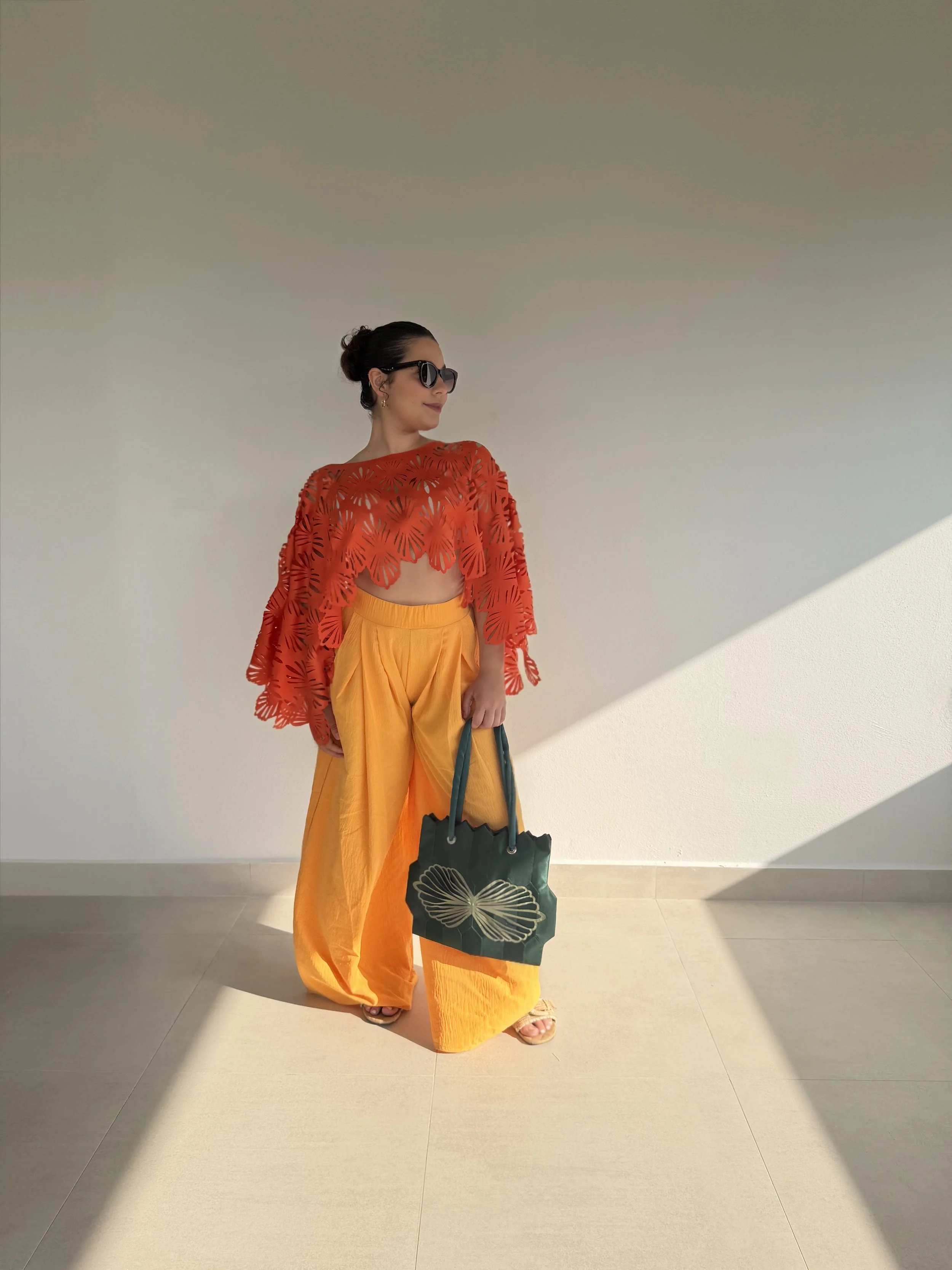 A woman wearing large black sunglasses, a red patterned top, and yellow wide-leg pants stands against a plain wall with dramatic shadows. She carries a dark green bag with a butterfly design, and wears open-toed sandals.