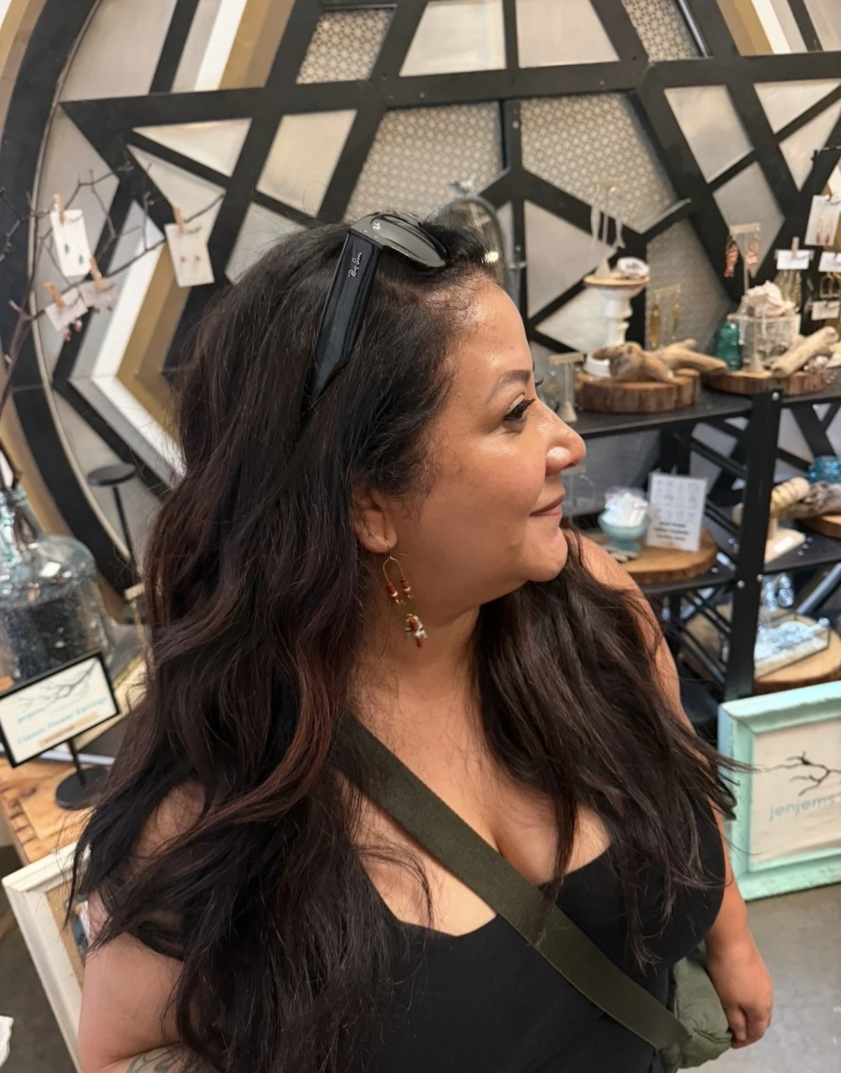 A woman with long dark hair and earrings standing in front of a black and beige geometric wall display of decorative items in a store.
