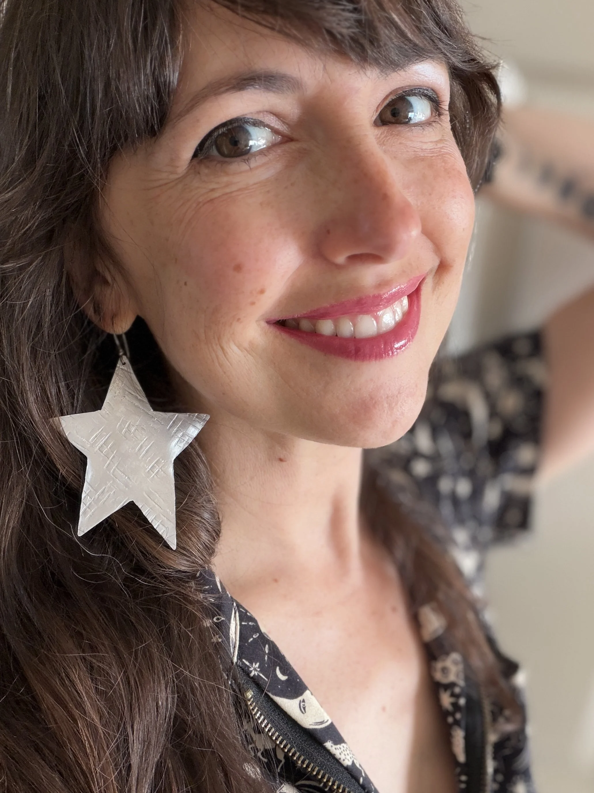 Close-up of a woman smiling, with long brown hair, wearing a black jacket with skull patterns and a white star-shaped accessory in her hair.