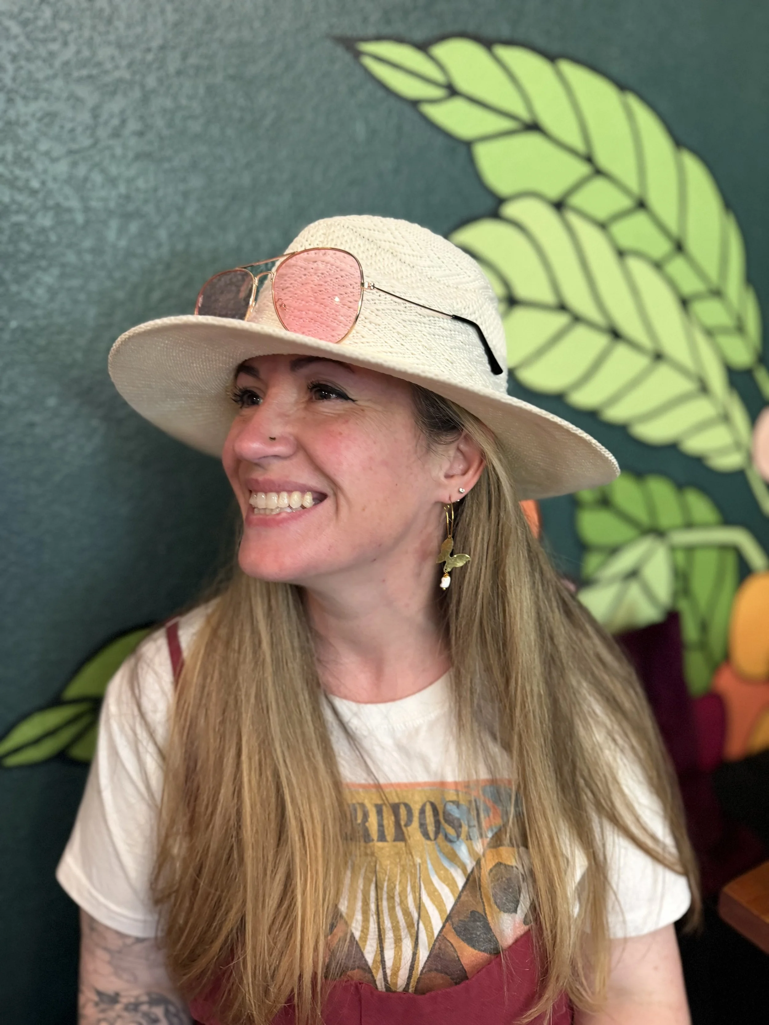 A woman smiling wearing a white wide-brimmed hat with pink sunglasses resting on it, and a graphic t-shirt with a red apron, standing in front of a dark green wall with large yellow-green leaf designs.