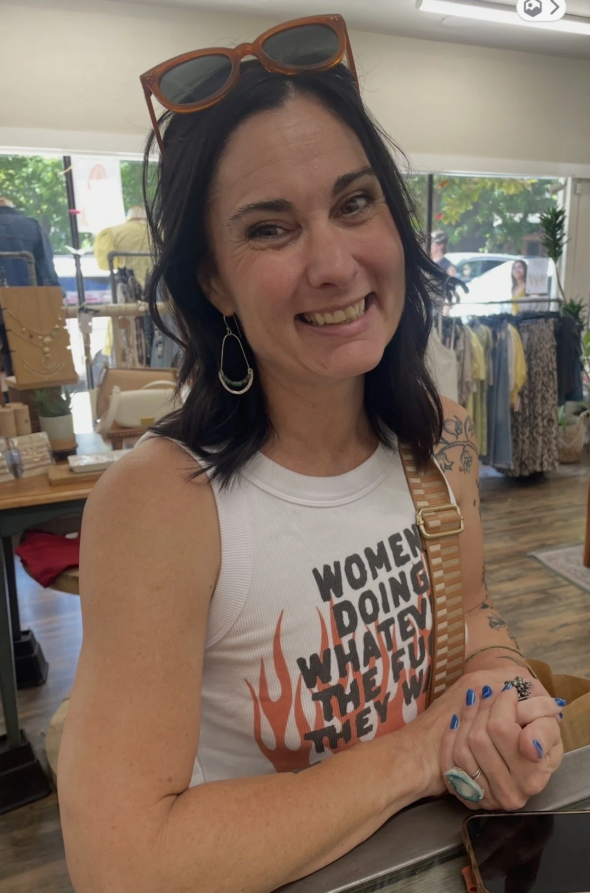 A woman with dark hair, sunglasses on her head, and earrings smiling inside a boutique store, wearing a white tank top with text, with clothing and accessories visible in the background.