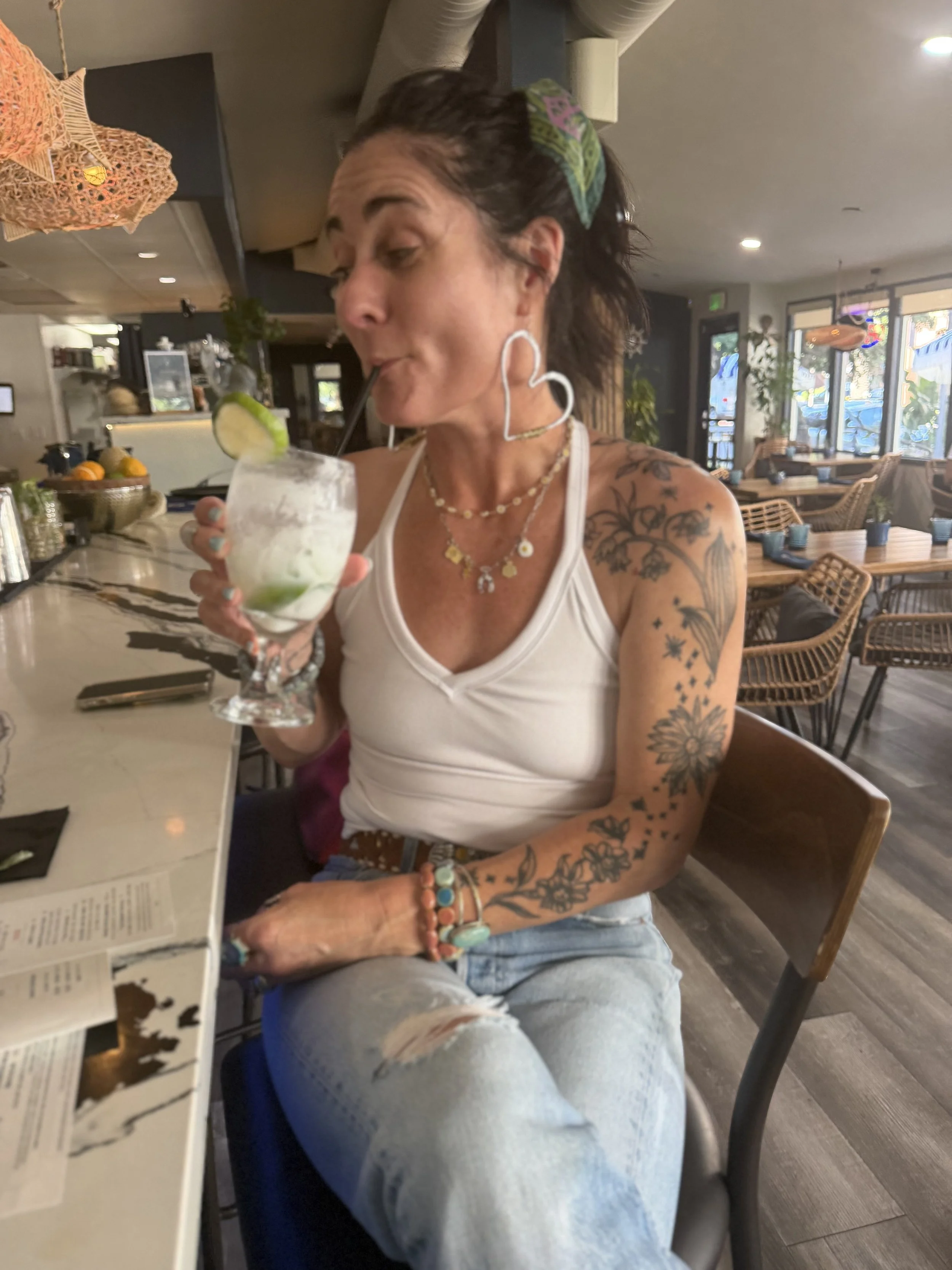 Woman with dark hair in a ponytail, wearing a white tank top, multiple necklaces, and tattoos on her arms, sitting at a restaurant table drinking a lime and lemon cocktail with a straw.