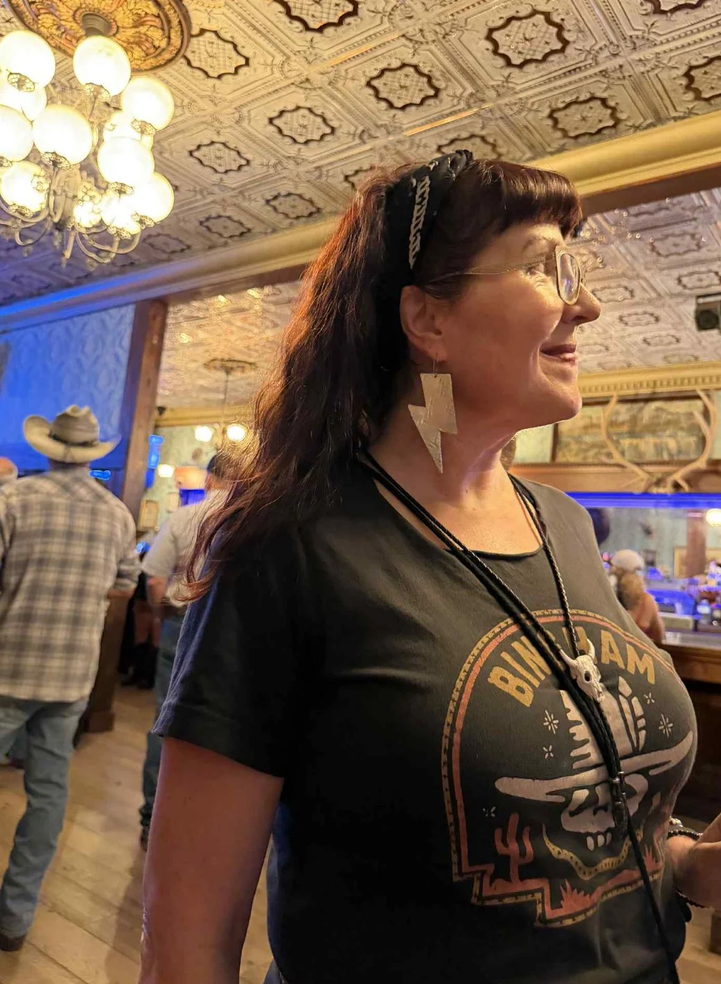 A woman with black hair, glasses, and lightning bolt earring standing inside a restaurant or bar, with a patterned ceiling, chandelier, and other patrons in the background.