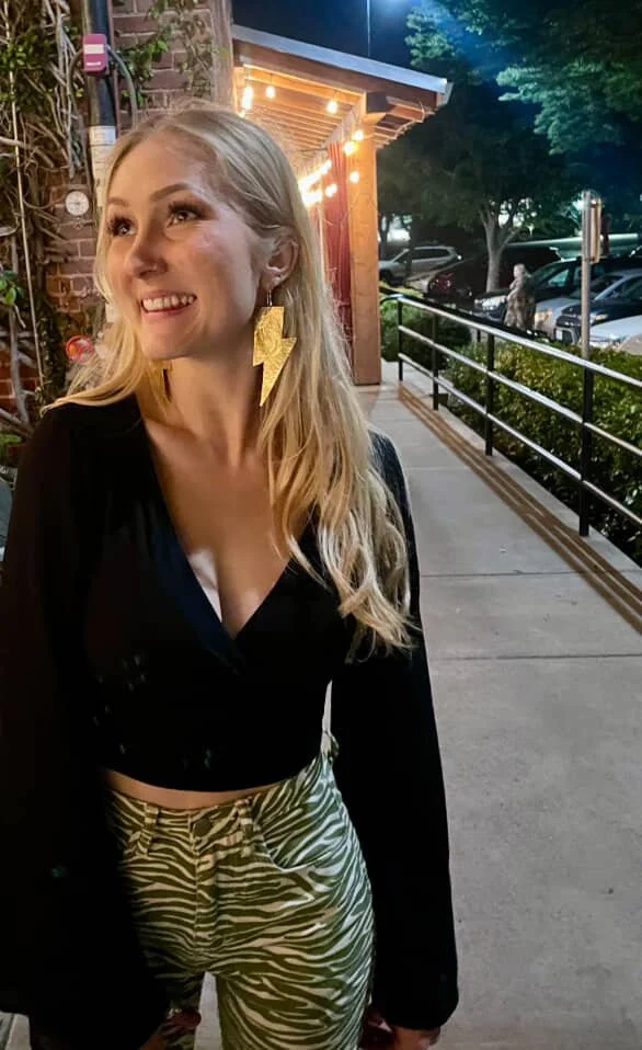 A young woman with long blonde hair smiling outdoors at night, wearing a black crop top, zebra-print pants, and large lightning bolt earrings, standing near a sidewalk with parked cars and trees in the background.